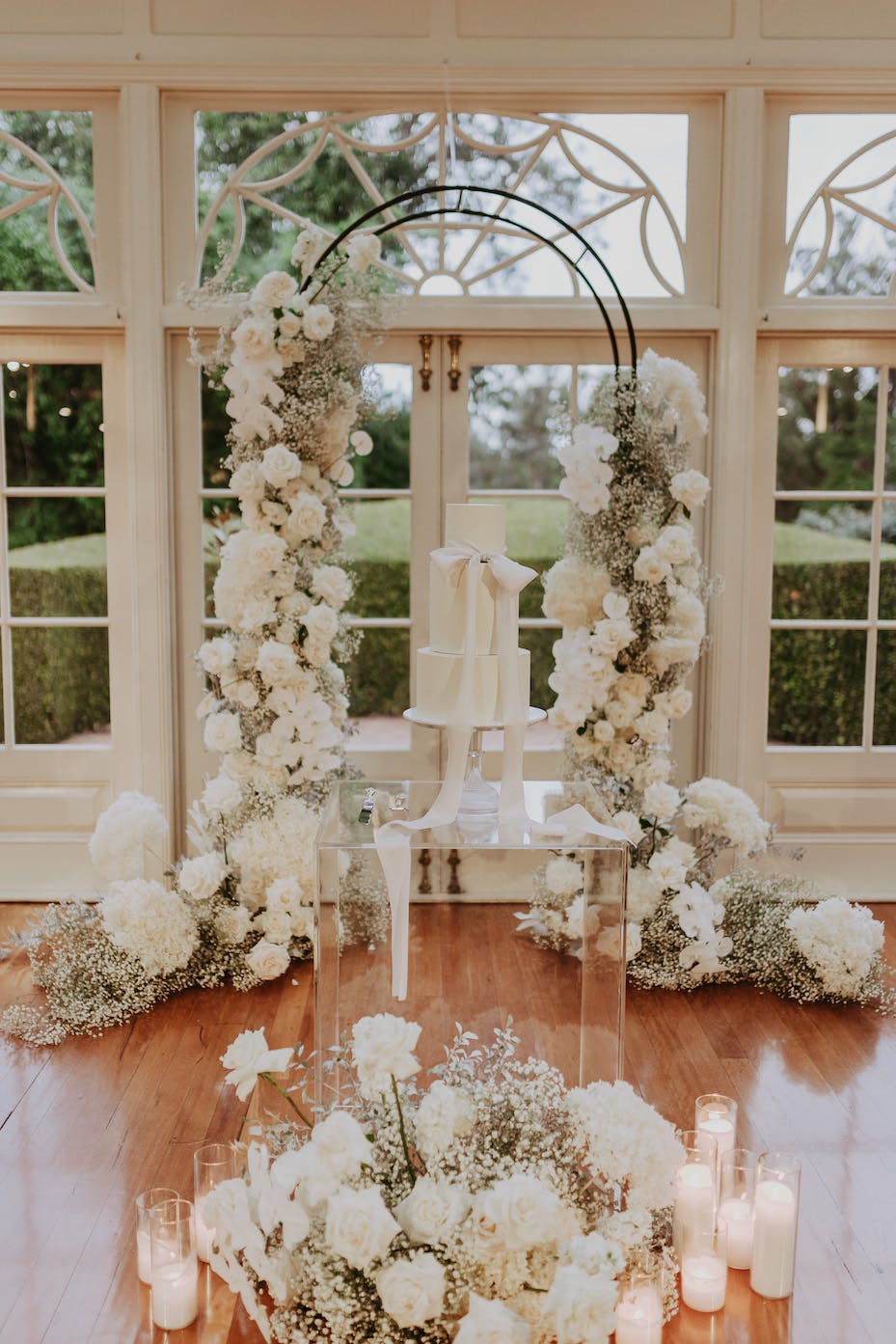 A wedding altar adorned with white and ivory flowers, including roses and baby's breath, arranged in tall, curved structures on both sides. A three-tiered white wedding cake sits on a glass stand in the center, with lit candles on the wooden floor.