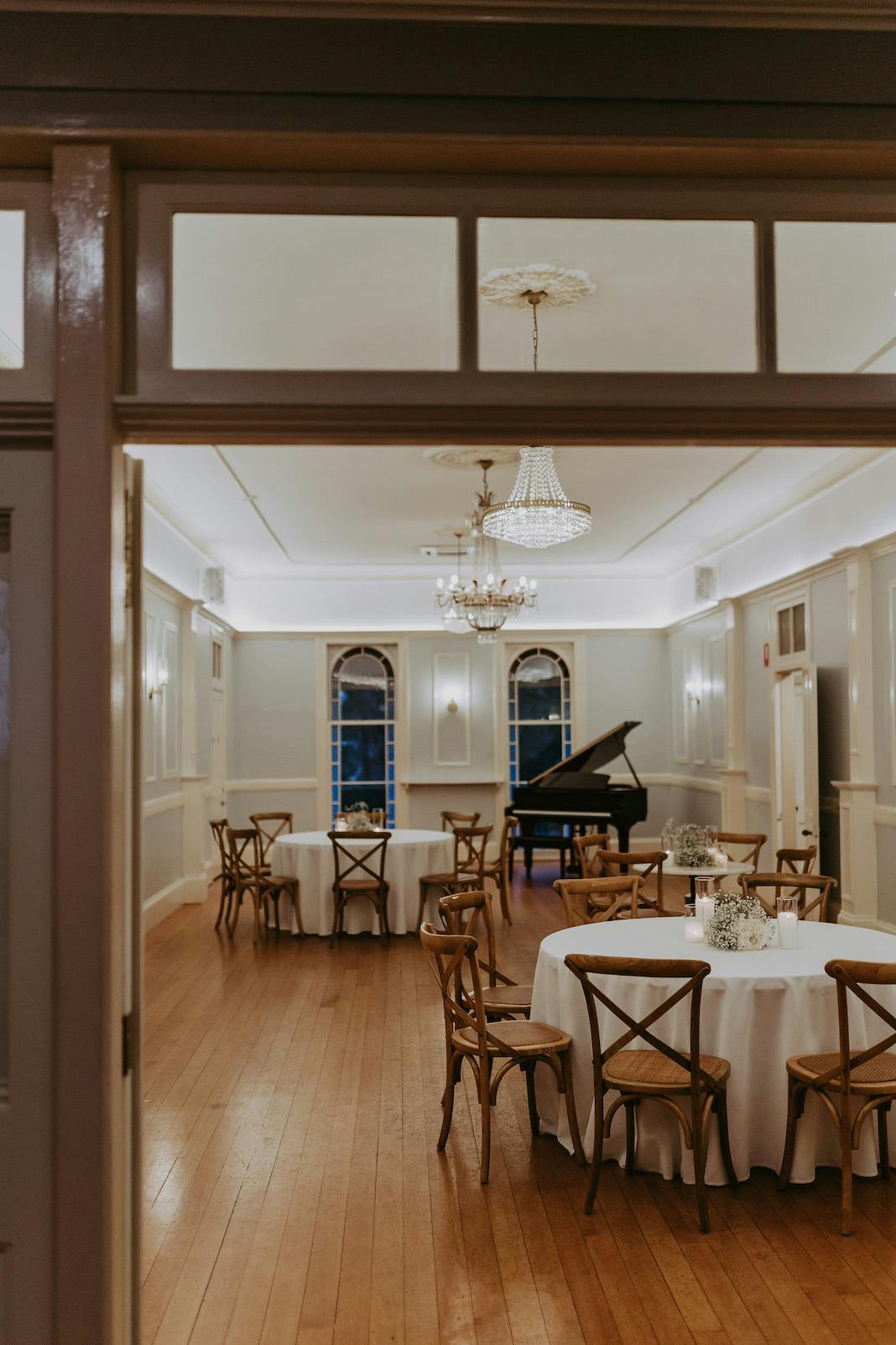 A spacious, elegantly decorated room with wooden floors features several round tables covered with white tablecloths and surrounded by wooden chairs. A black grand piano sits at the back of the room near large arched windows, and crystal chandeliers hang from the ceiling.