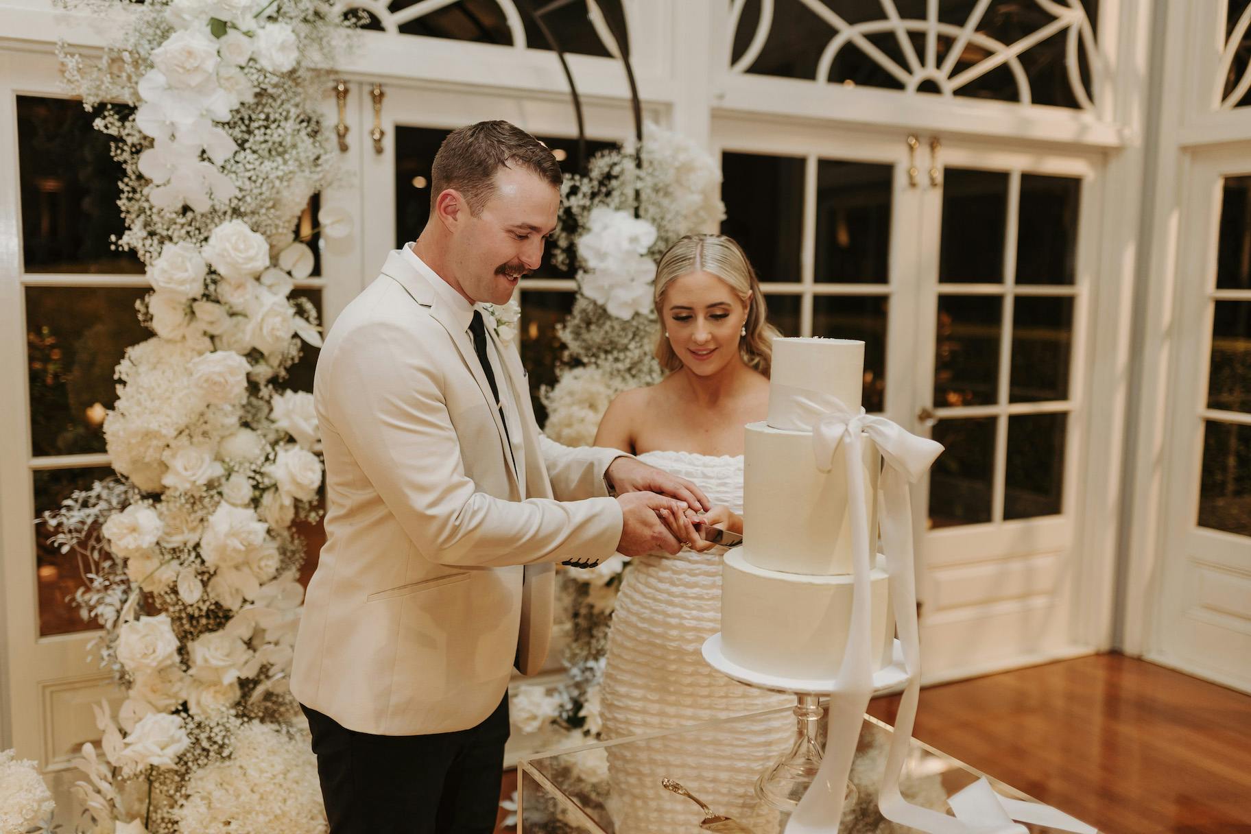 A bride and groom, both in white attire, stand in front of a tall, white wedding cake adorned with flowers and ribbons. The groom holds a cake knife while the bride touches his hand, both smiling as they prepare to cut the cake. A floral arch frames them in the background.
