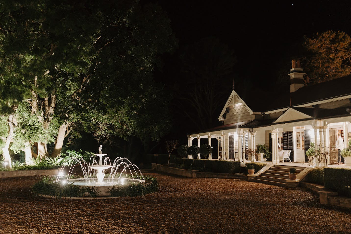 A nighttime scene of a well-lit, large house with a wraparound porch and multiple windows. In the yard, there is a decorative lit-up fountain surrounded by gravel pathways. Tall trees frame the left side, casting shadows on the building and surroundings.