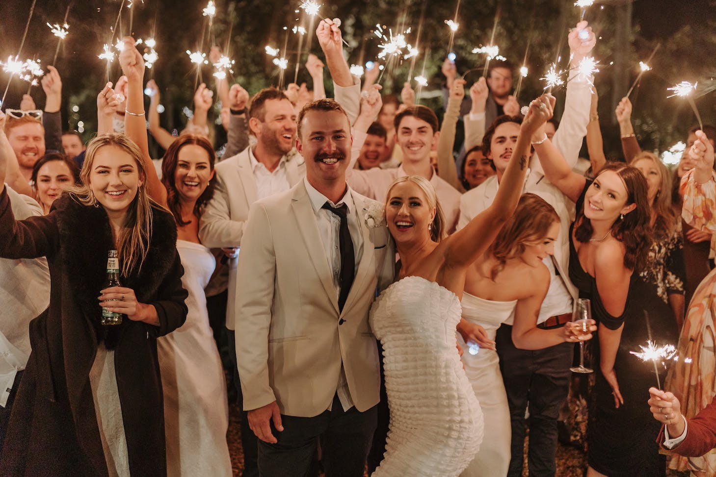 A joyful bride and groom are surrounded by friends and family holding sparklers at an outdoor celebration. The bride is in a white strapless dress, and the groom is in a light-colored suit with a black tie. Everyone is smiling, laughing, and enjoying the festive atmosphere.