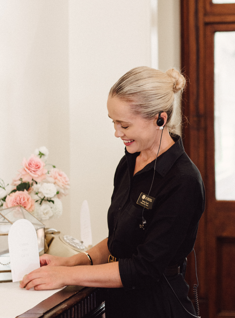 A woman with blonde hair tied in a bun, wearing a black uniform and a headset, stands behind a counter smiling and holding a small document. Behind her are pink and white flowers in a vase. The setting appears to be a reception or customer service desk.