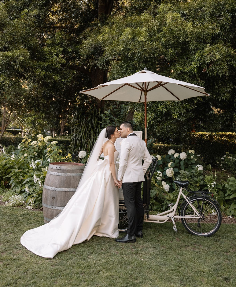 A newlywed couple stands holding hands and kissing outdoors in a lush garden. The bride wears a long, flowing wedding dress with a veil, and the groom is in a light-colored suit. They're by a bicycle with an umbrella and a wooden barrel nearby, surrounded by greenery and flowers.