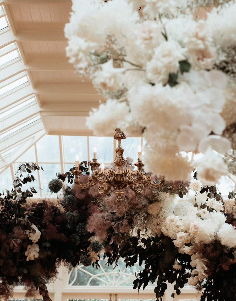 A luxurious chandelier with intricate golden details hangs from a high, glass-paneled ceiling. Surrounding the chandelier are lush, cascading arrangements of white and pastel flowers, creating an ethereal and elegant atmosphere in the room.