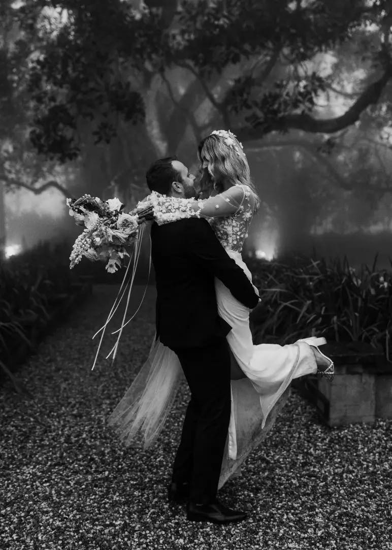 A black-and-white photo depicting a groom lifting his bride in his arms. The bride is holding a bridal bouquet and wearing a flowing wedding gown with veil, while the groom is in a dark suit. They are outdoors, surrounded by trees and mist, creating a dreamy atmosphere.