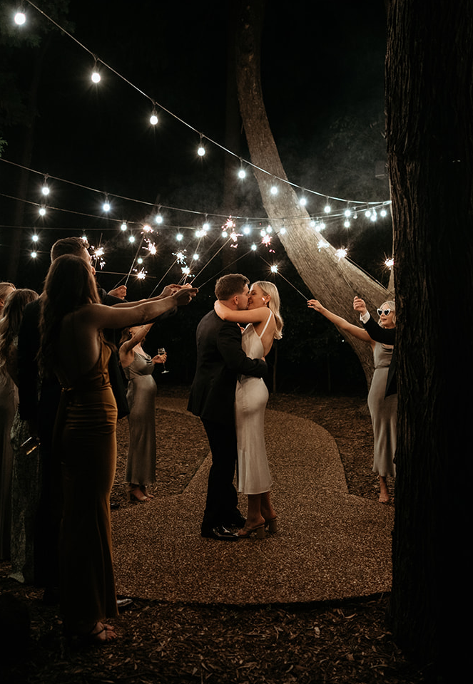A couple shares a kiss under string lights during a nighttime outdoor wedding ceremony. Guests surround them holding sparklers, creating a festive and romantic atmosphere. The scene is illuminated by the warm glow of the lights and sparklers.