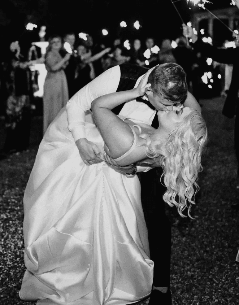A couple shares a passionate kiss during their wedding dance at night. The groom dips the bride, who is wearing a flowing white dress. Guests holding sparklers surround them, adding a magical glow to the scene.