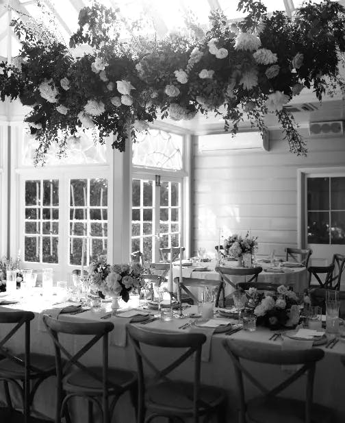 a black and white photograph of the wedding venue set up ready for the wedding breakfast, beautiful flowers hang from the ceiling and also form table decorations.