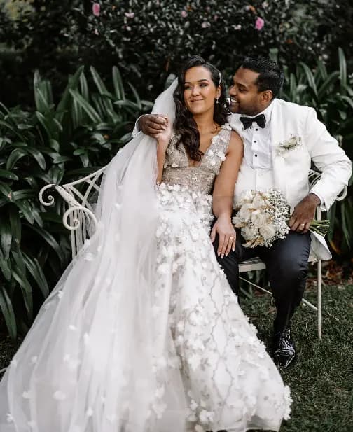 A bride and groom sit on a white bench in a garden. The bride, in a floral lace gown and long veil, holds a bouquet of white flowers. The groom, in a white tuxedo jacket with black pants and bow tie, drapes his arm over the bride's shoulder as they smile.
