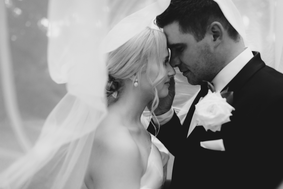 A black and white photo of a bride and groom embracing under a veil. The bride has her hair in an updo and is wearing a strapless dress with pearl earrings. The groom, in a suit with a white rose boutonniere, gently touches the bride's cheek, their foreheads touching.