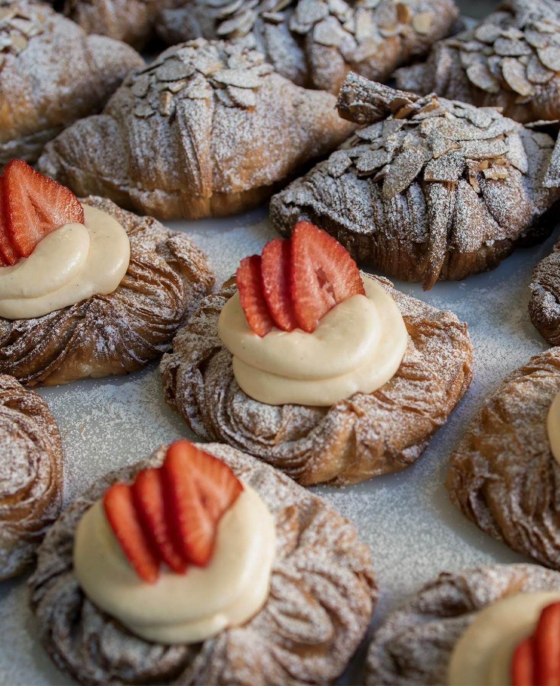 A tray filled with freshly baked pastries. Some pastries are topped with powdered sugar and almond slices, while others have a swirl of cream and sliced strawberries on top. The pastries have a golden, flaky texture and are arranged closely together.