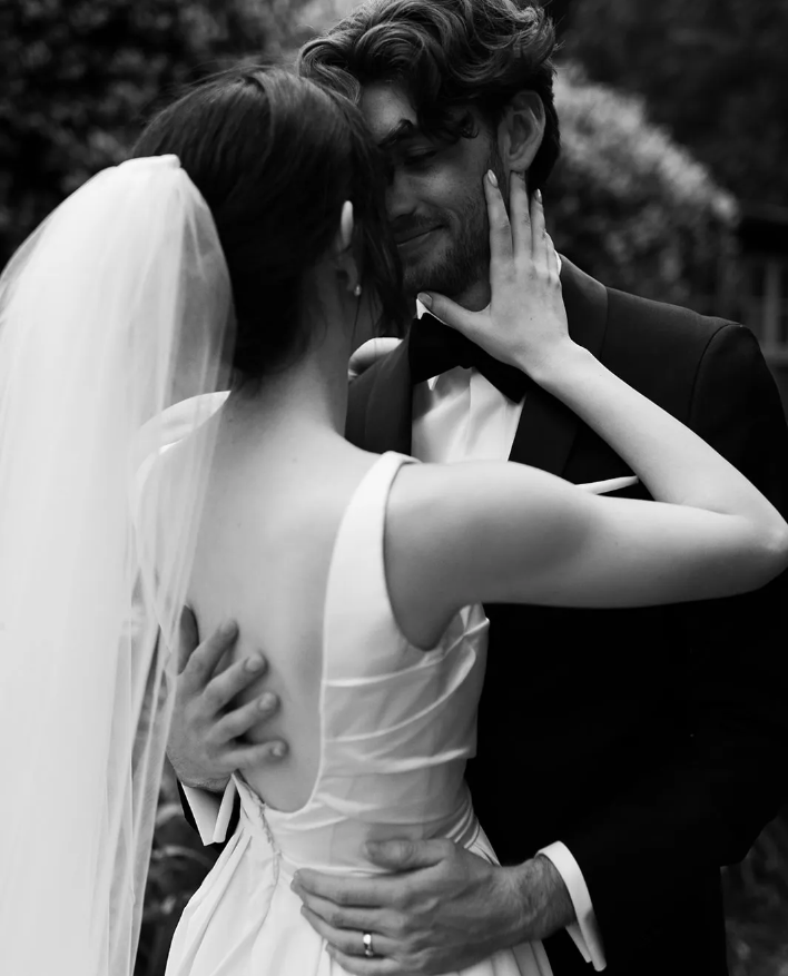 A black and white image of a bride and groom. The bride is wearing a veil and a white dress with a low back, and she gently holds the groom's face with one hand. The groom is dressed in a suit and bow tie, and they are gazing into each other's eyes lovingly.