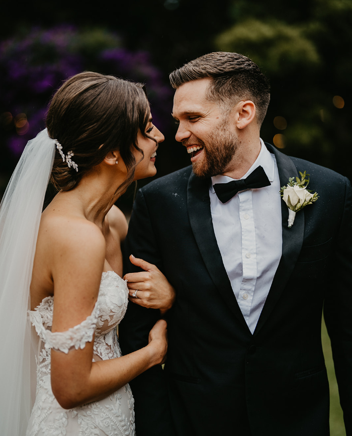 A bride and groom smile joyfully while looking at each other. The bride wears a white lace dress with off-the-shoulder sleeves and a long veil, while the groom is dressed in a black tuxedo with a white bowtie and boutonnière. The background is blurred and dark.