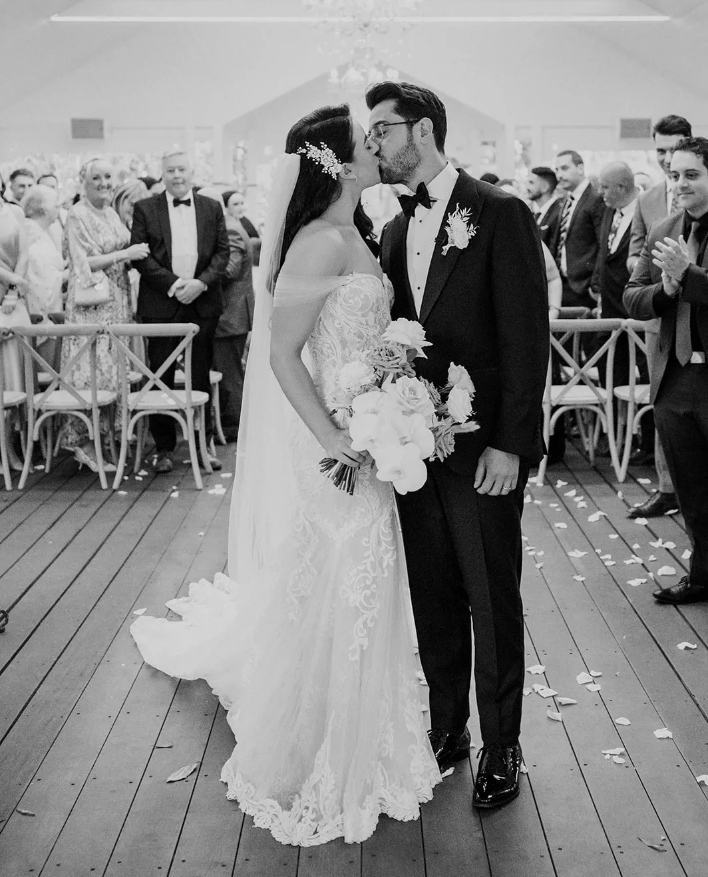 A bride and groom share a kiss at their wedding ceremony. The bride is wearing an off-the-shoulder lace gown and holding a bouquet of flowers, while the groom is in a classic tuxedo. Guests stand and applaud in the background, and rose petals are scattered on the floor.