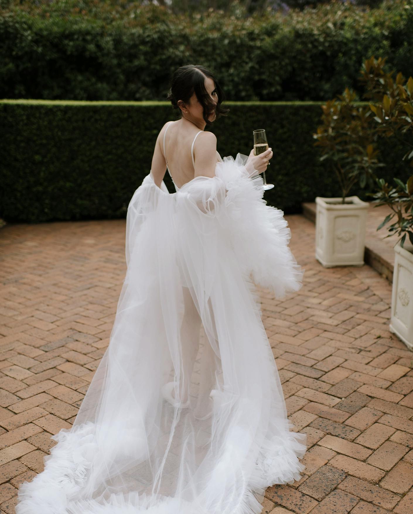 A woman in a flowing white wedding dress with a long train stands on a brick path, holding a champagne glass. She is turned away from the camera, and the scene is set against a backdrop of greenery.