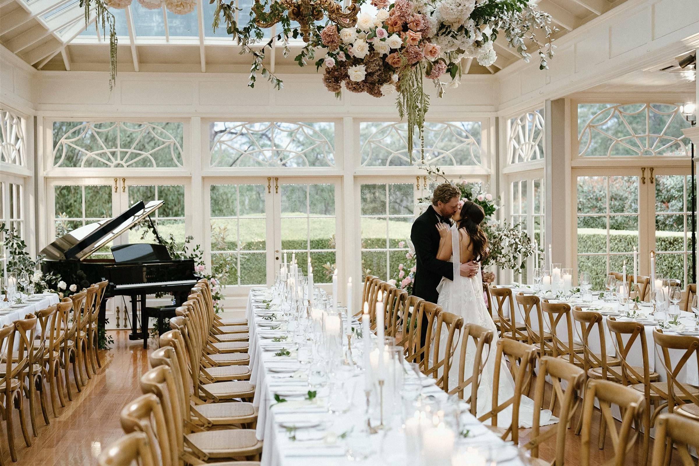 A couple in wedding attire share a kiss in an elegant, bright reception hall. The room is decorated with a hanging floral arrangement and long dining tables set for guests. Large windows overlook a lush garden, and a grand piano is positioned in one corner.