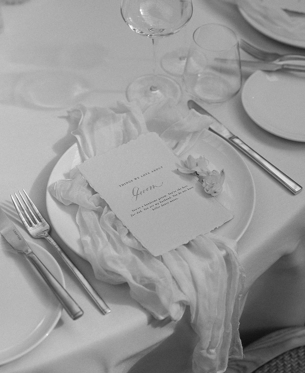 A black and white image of a table setting with a plate, a menu card, cutlery, glasses, and a decorative cloth. The plate holds a card that reads "Here's to love, honor," with elegant cursive handwriting. A flower-like decoration is placed beside the card.