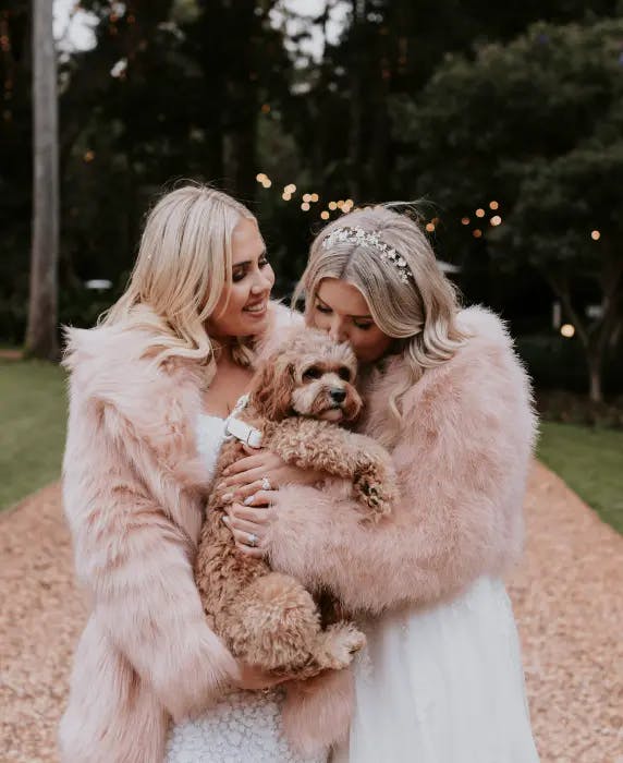 Two women in white dresses and pink fur coats smile while one holds a small, curly-haired dog. They are standing on a gravel path surrounded by trees with string lights in the background, creating a warm and intimate setting.