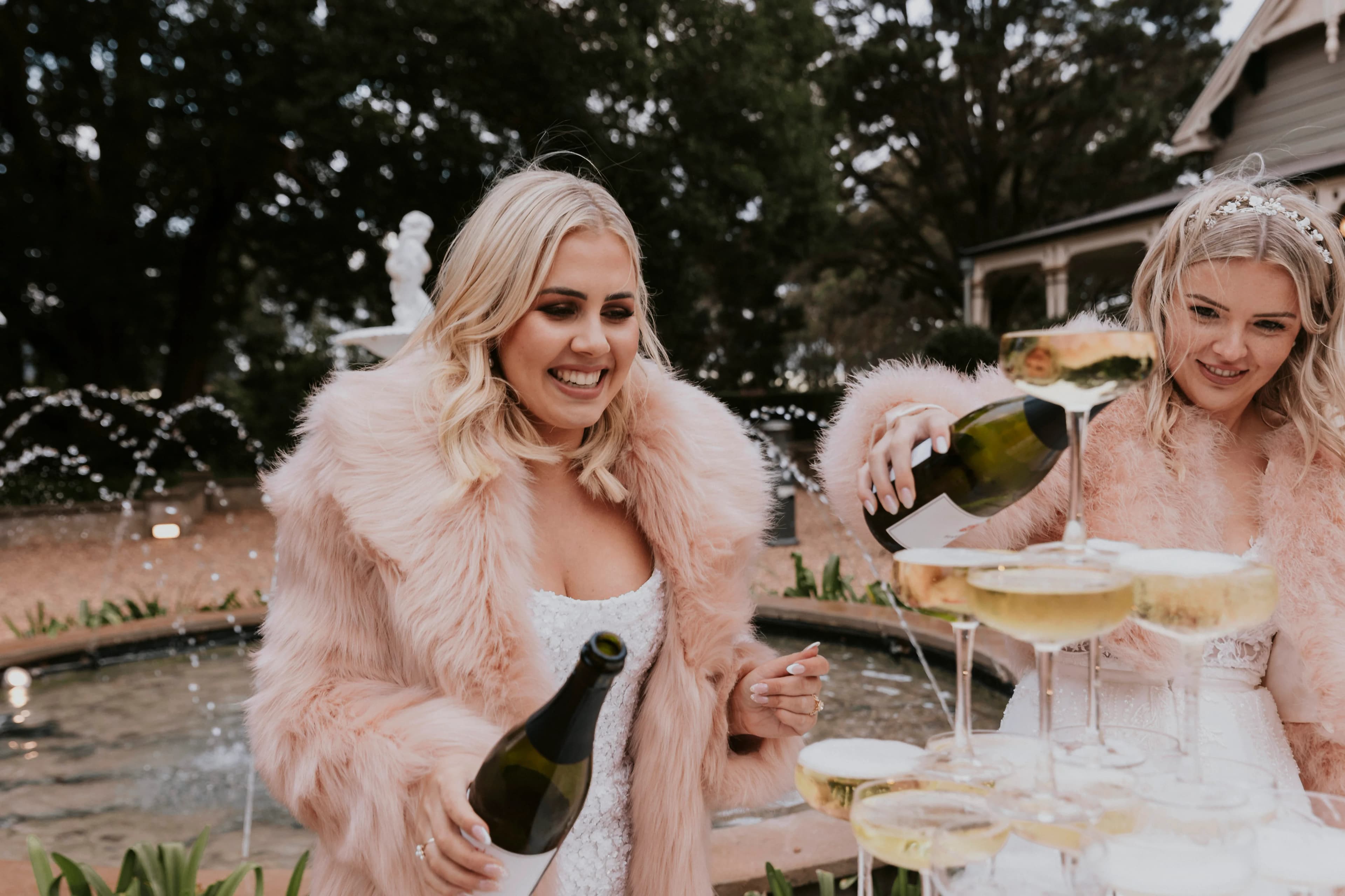 Two smiling women in fur coats are pouring champagne into a tower of glasses outdoors. A stone fountain and greenery are visible in the background. One woman has platinum blonde hair and is pouring champagne on the left, while the other woman is pouring on the right.