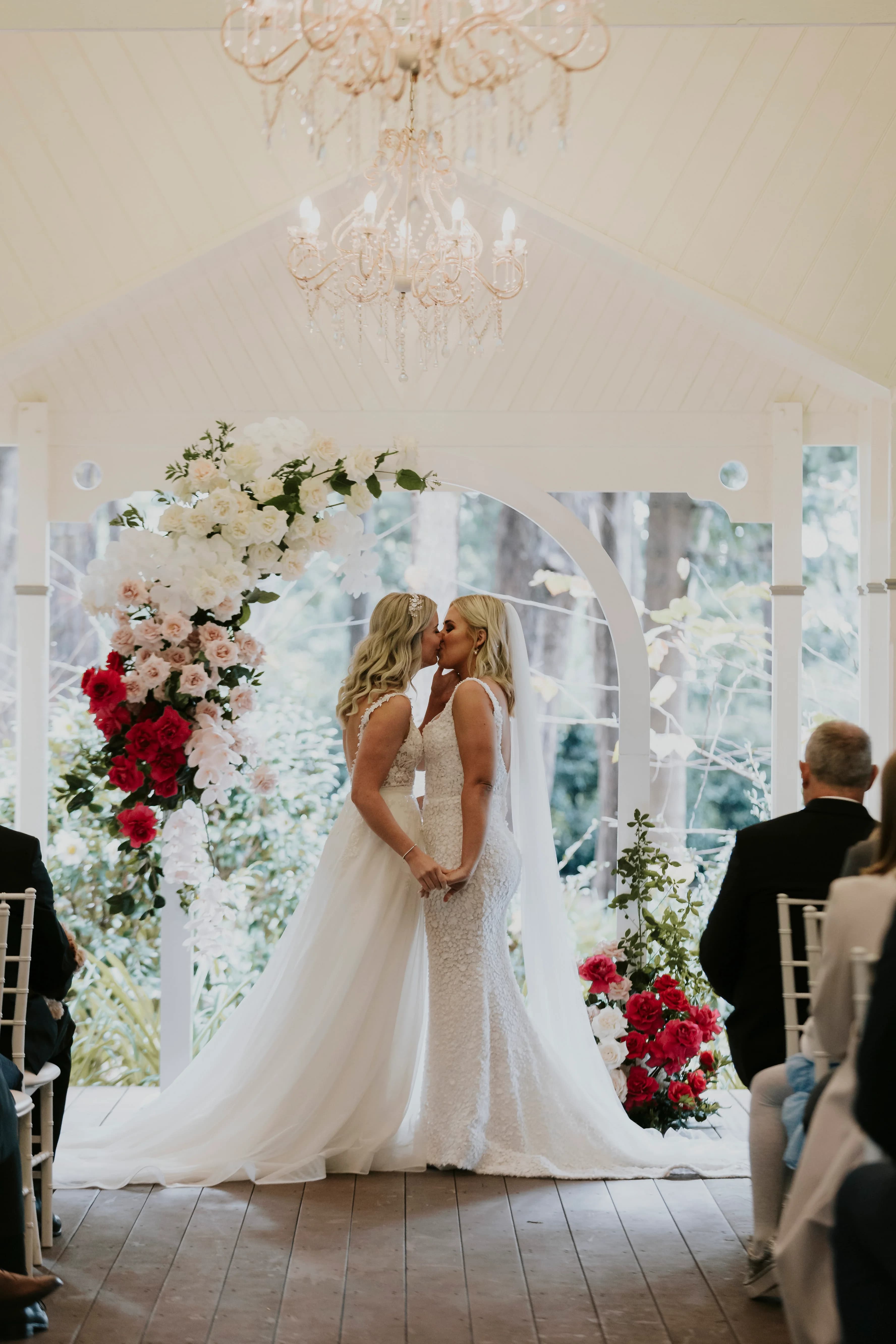 Two brides share a kiss under a floral arch at their wedding. Both are in white gowns; one has a strapless dress and the other a dress with cap sleeves. They stand in an outdoor setting, surrounded by guests seated on white chairs. A chandelier hangs above them.