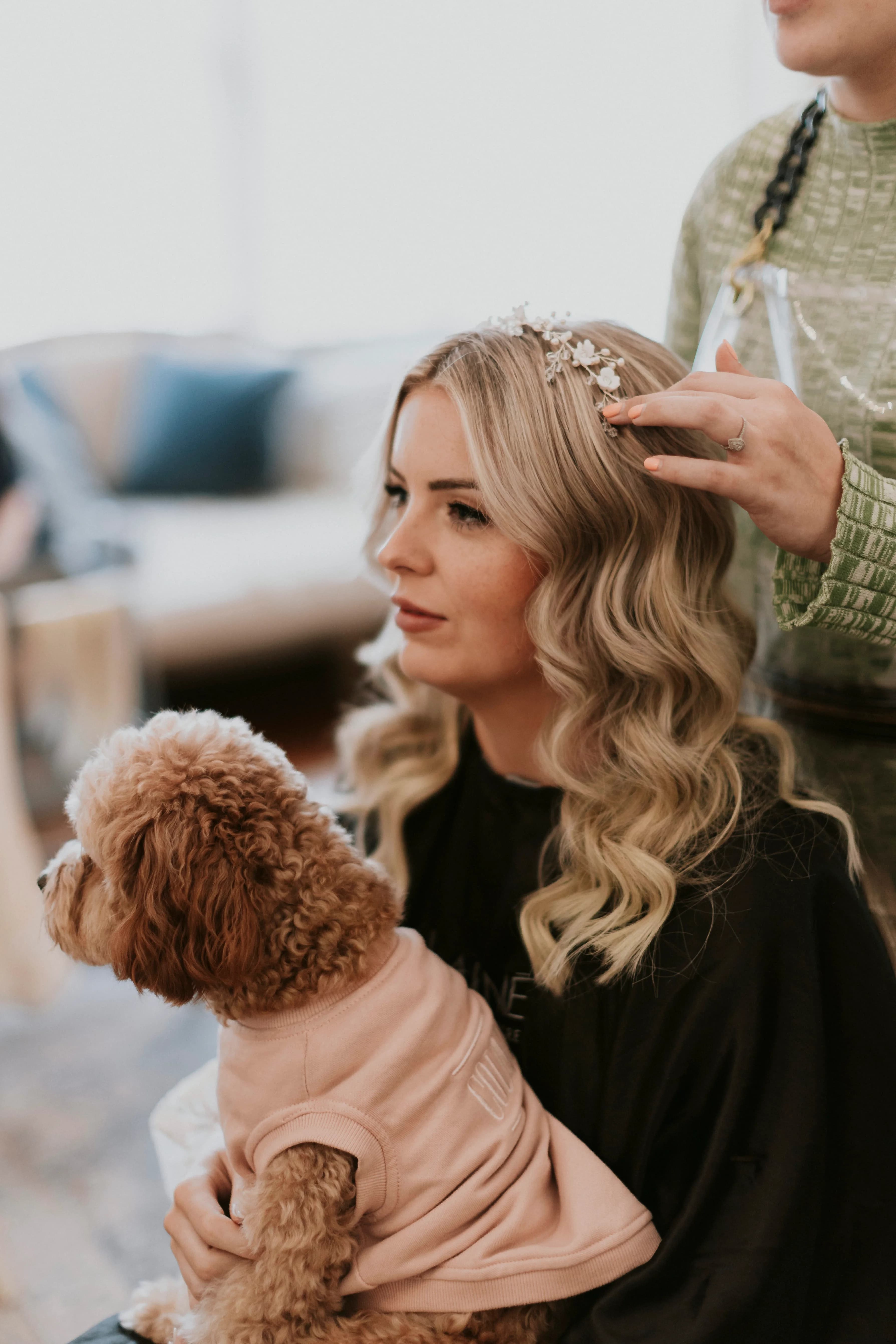 A woman with wavy blonde hair is sitting while someone styles her hair, adding a floral hair accessory. She is holding a small, curly-haired dog wearing a pink sweater on her lap. The background features a blurred living room setting with a sofa and pillows.