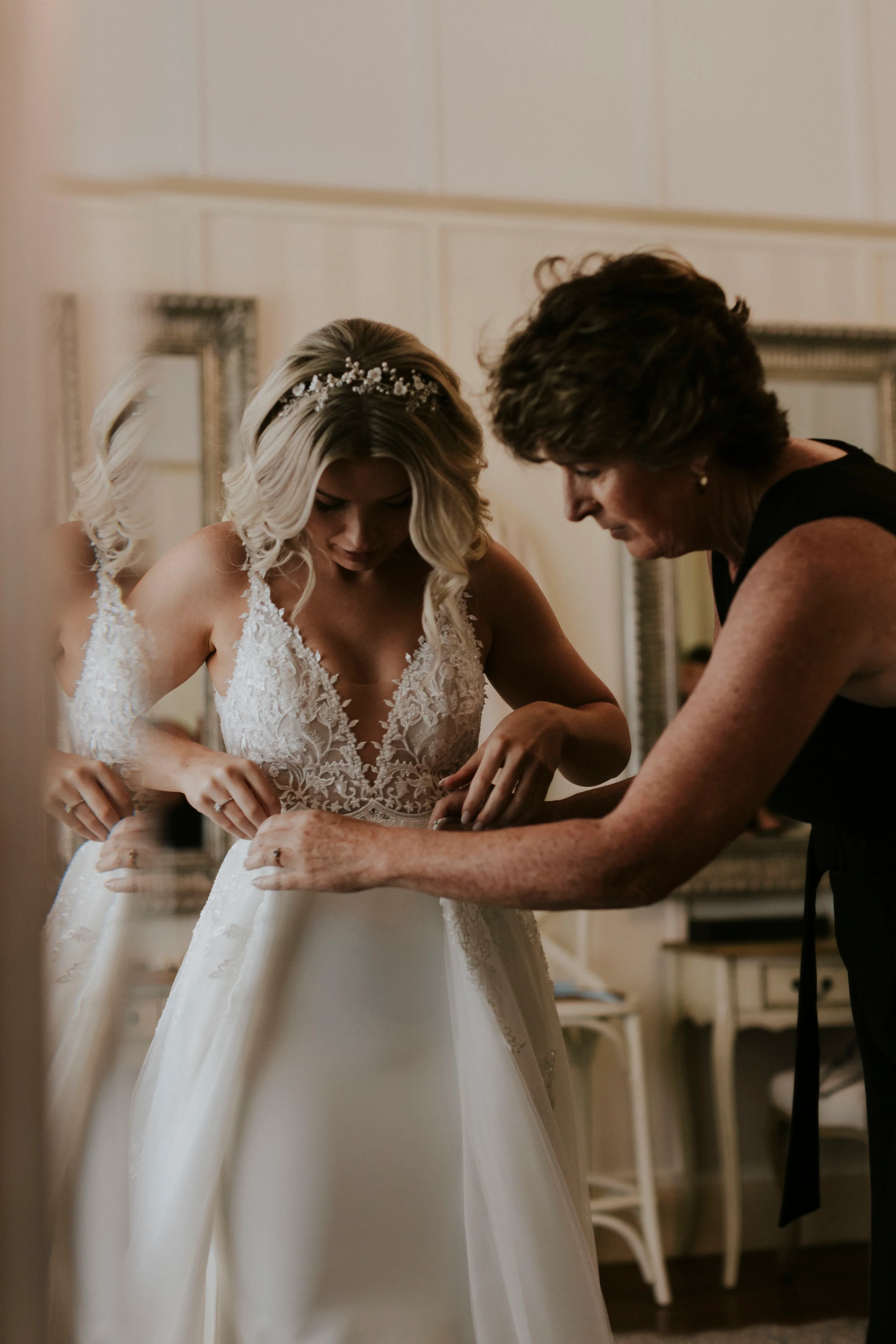 A bride in a white lace wedding gown, with a headpiece adorning her wavy blonde hair, stands with an older woman who is assisting her with the dress. They are in a room with mirrors and light-colored walls. The atmosphere appears calm and focused.