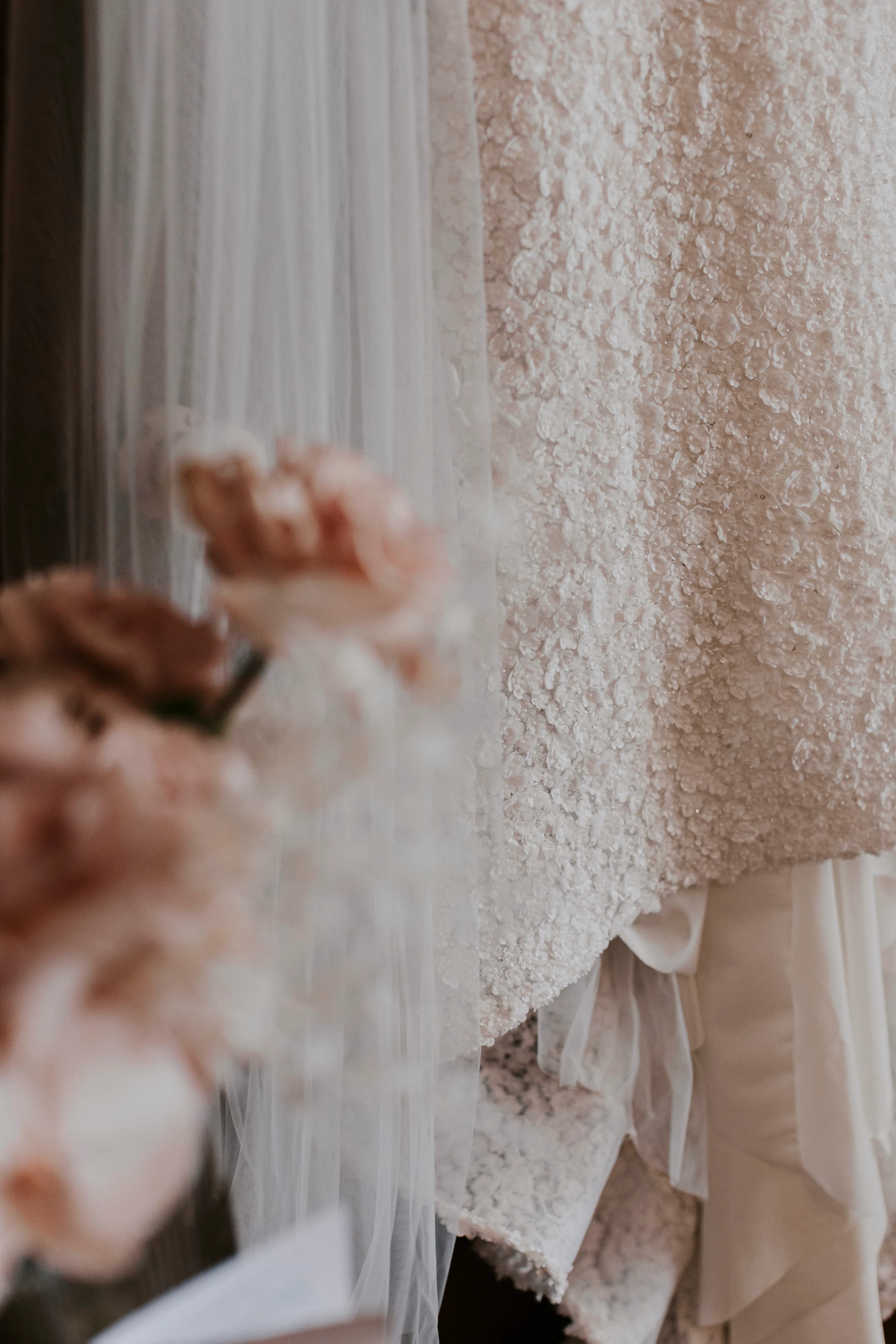 Close-up of a textured bridal gown with layers of white fabric and a sheer veil. Blush-colored flowers in soft focus are in the foreground, creating a romantic and elegant atmosphere.