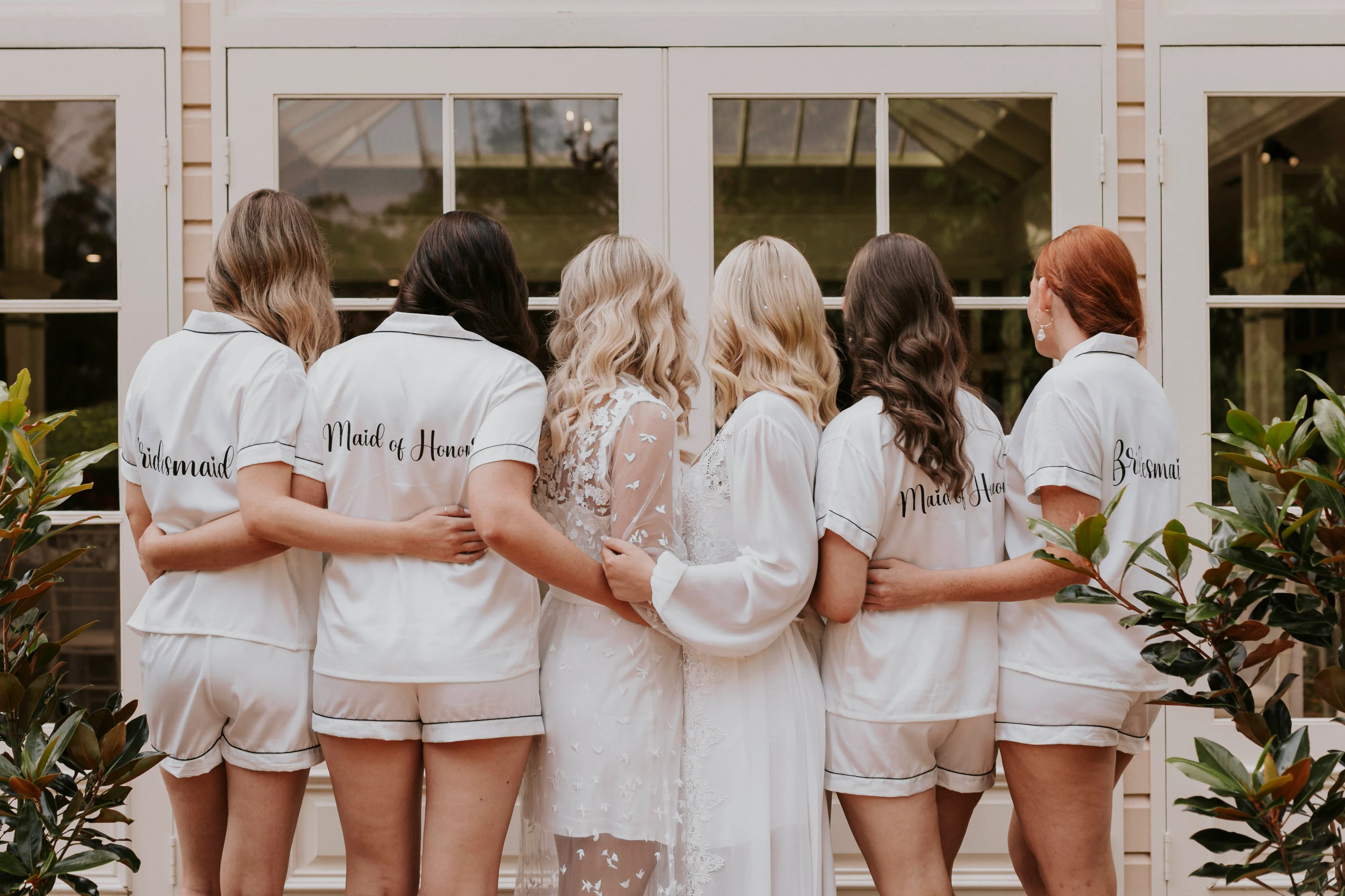 A group of six women, seen from behind, stand arm-in-arm in front of a window. The second woman from the left wears a sheer white robe, while the others wear matching white pajamas with titles like "Maid of Honor" and "Bridesmaid" written on the back.