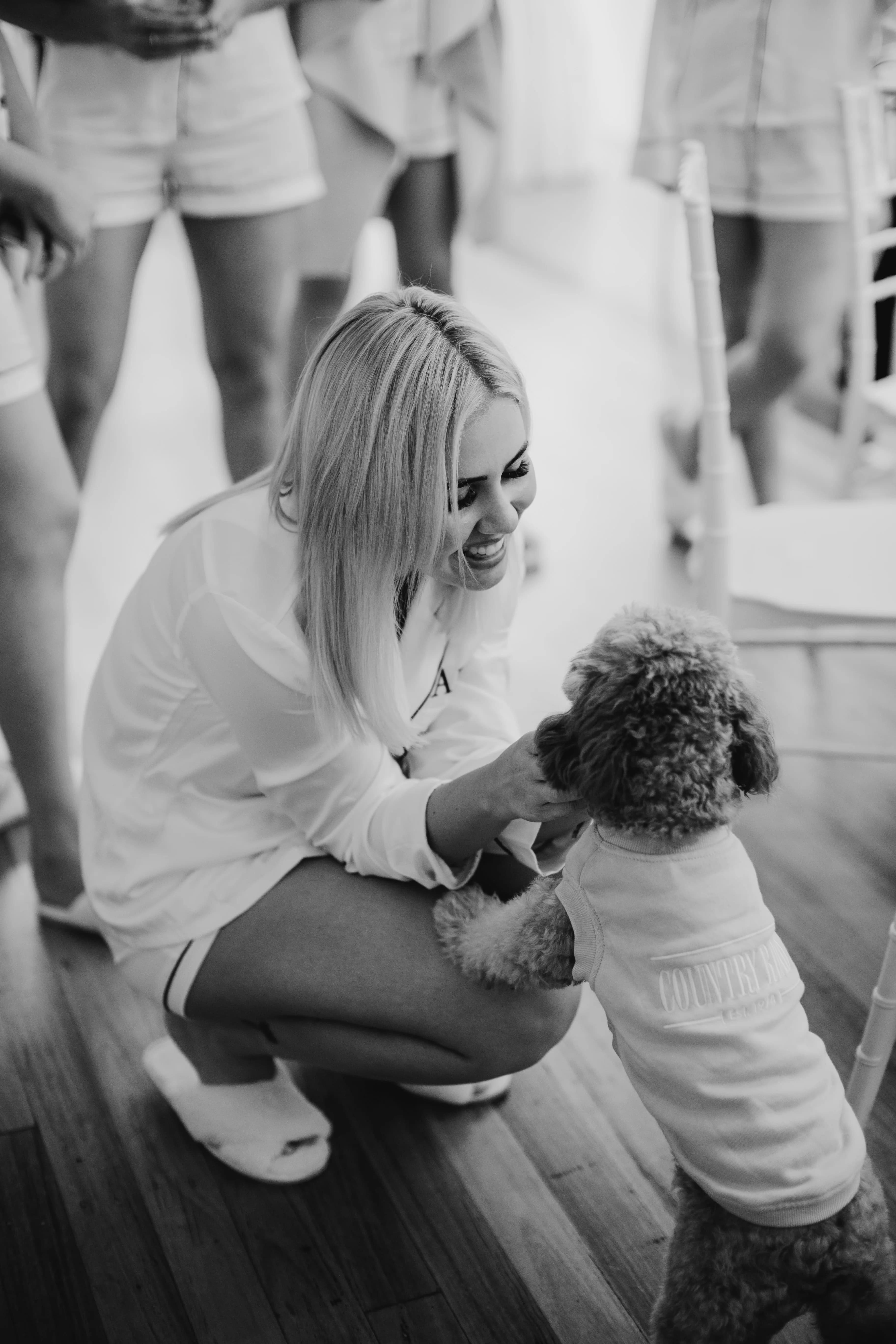 Black and white image of a woman with long blonde hair, wearing a shirt and shorts, kneeling and smiling as she interacts with a small dog on its hind legs. The dog is dressed in a cute outfit. Other people, dressed casually, are partially visible in the background.