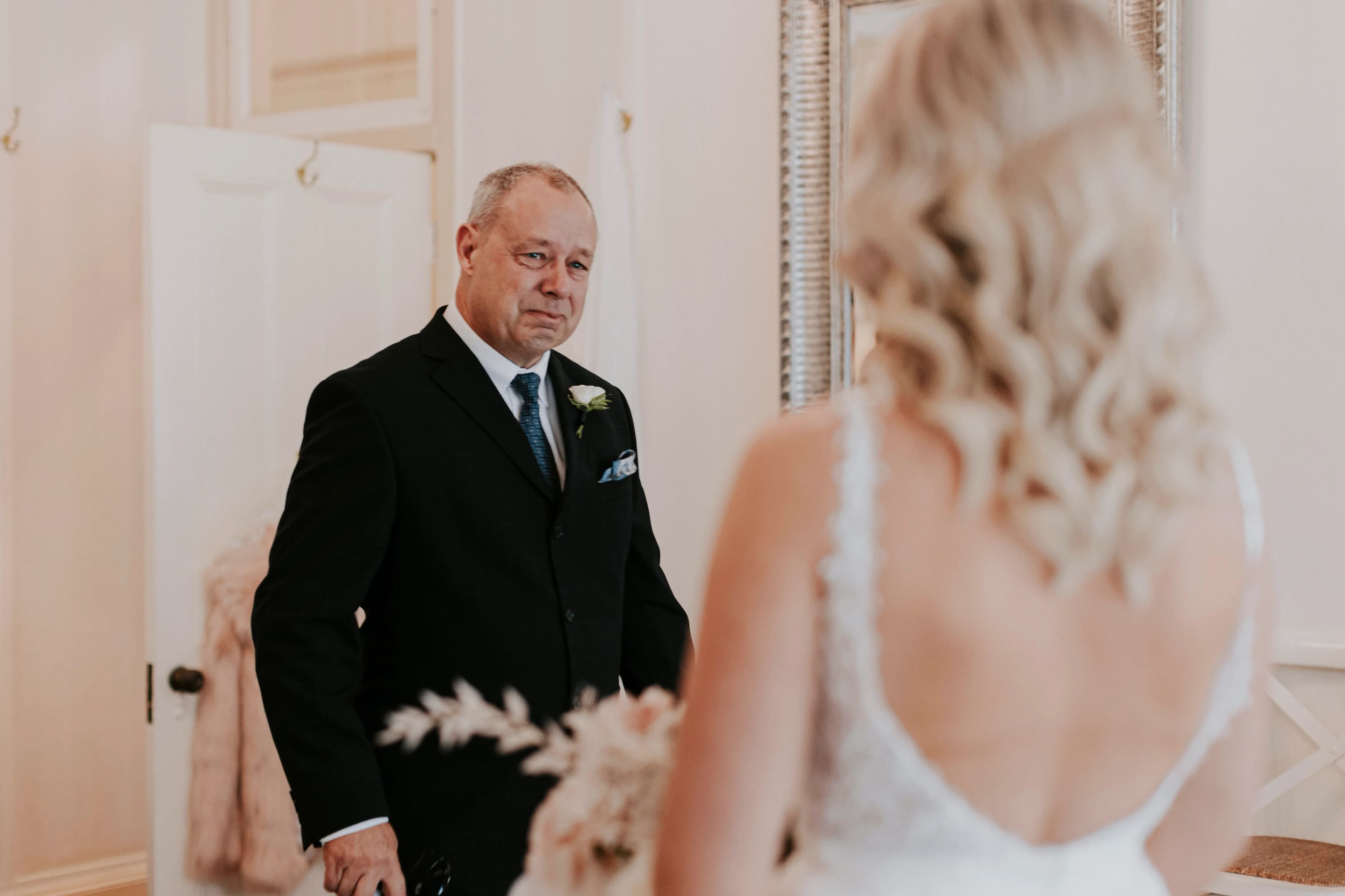 A man in a suit and tie stands in a room looking emotionally at a woman in a white dress with her back to the camera. The woman appears to be a bride, with long, wavy blonde hair and a bouquet. The room is decorated simply with light colors.