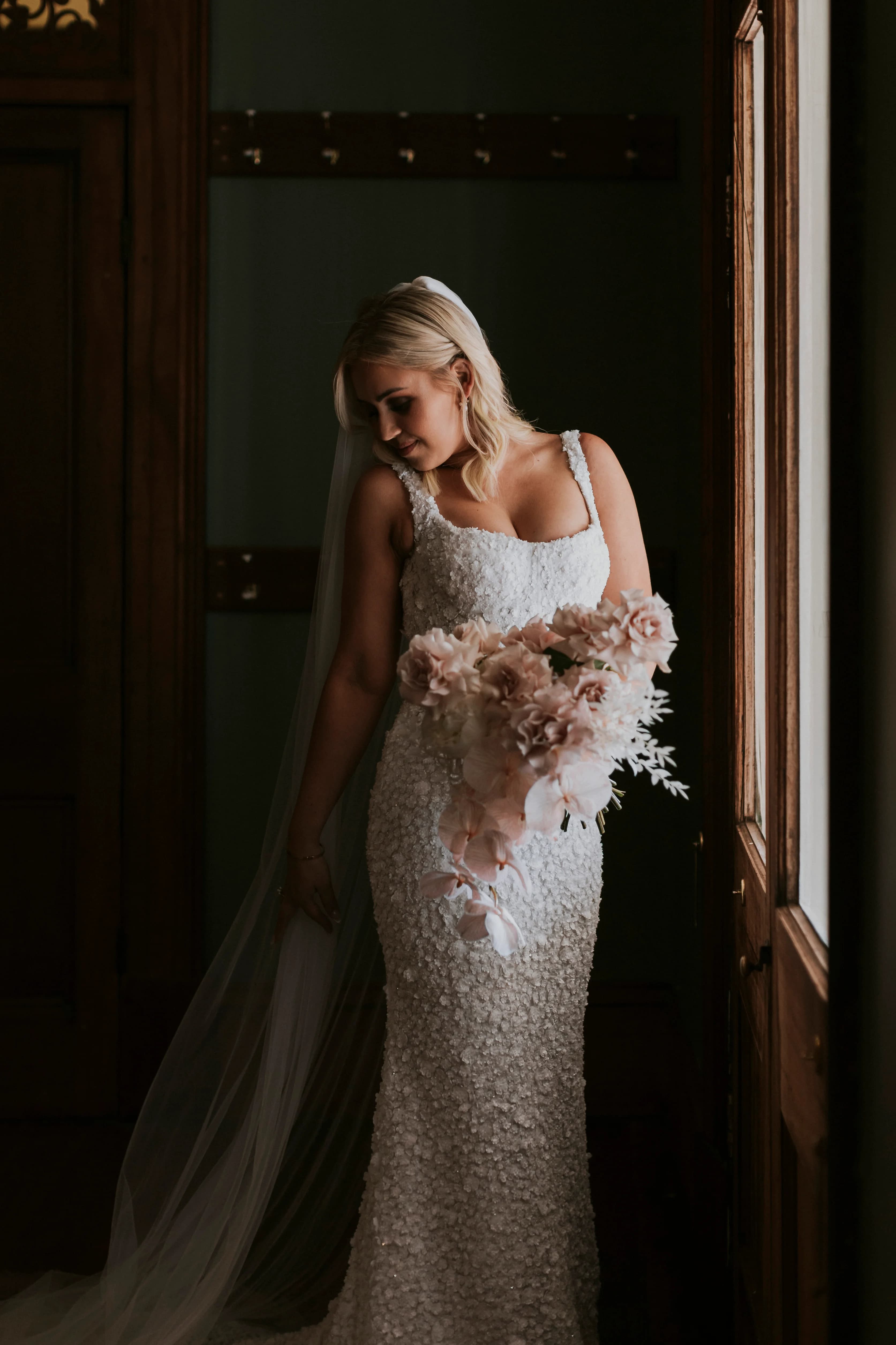 A bride in a white, textured gown holding a large bouquet of pale pink flowers stands near a wooden door. The light from outside softly illuminates her face and dress, highlighting the intricate details of the fabric and the flow of her veil down her back.