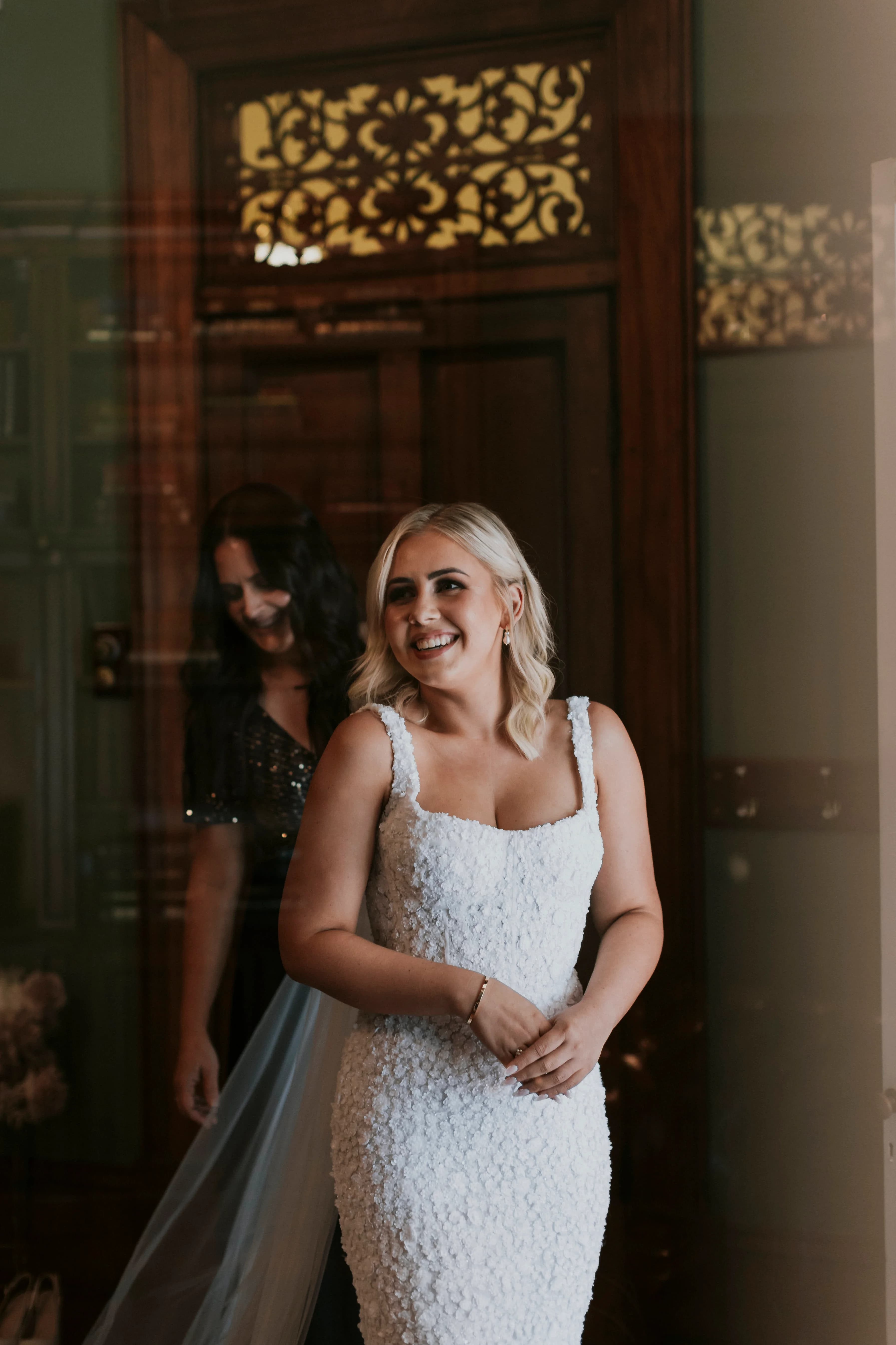 A bride in a white, textured wedding dress and short blonde hair smiles joyfully, standing indoors. A woman in a dark dress stands behind her, partially visible. The room features ornate wooden doors with intricate designs on the glass panels.