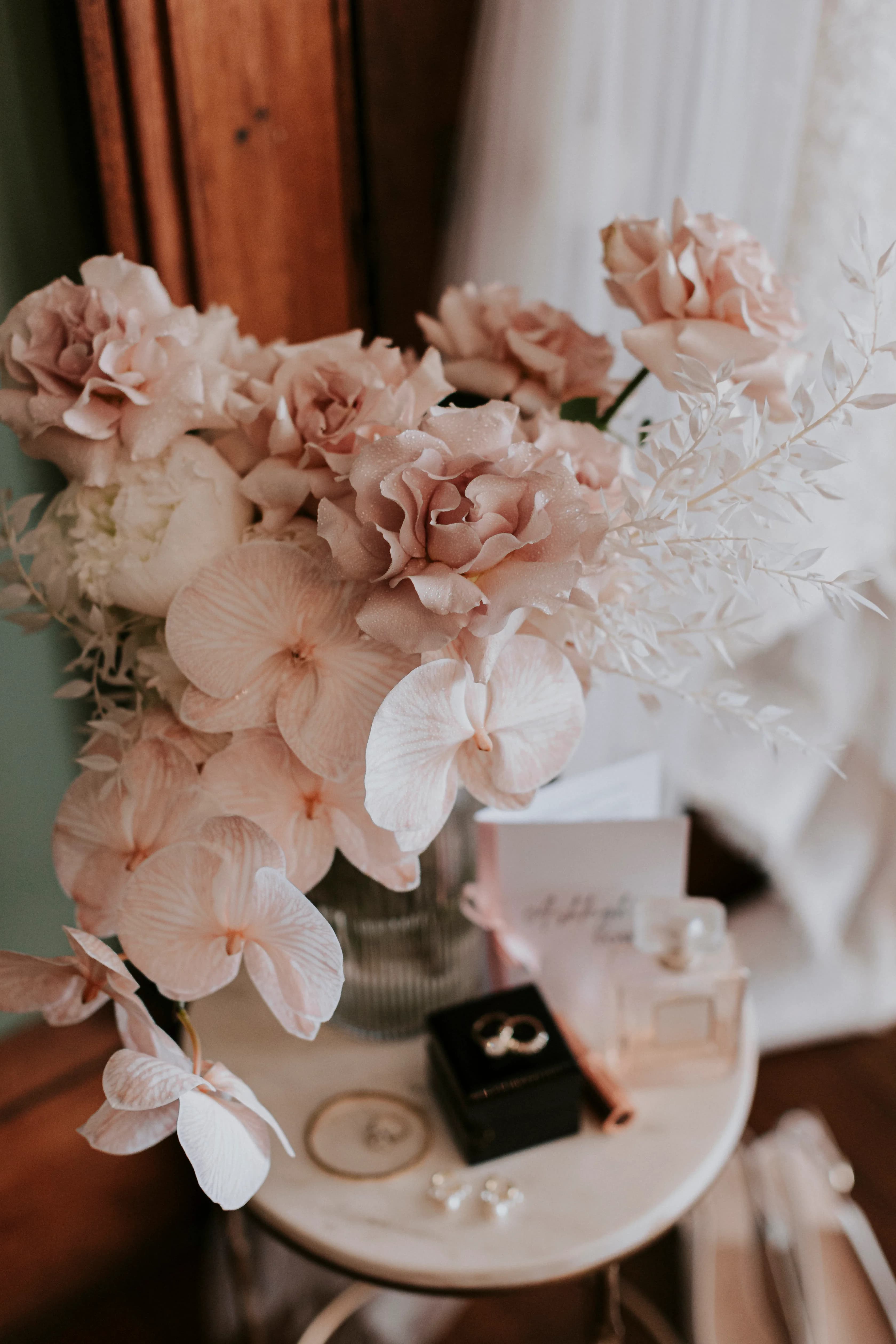 A close-up of a bouquet with blush and ivory flowers, including roses and orchids, in a vase on a round table. Beside the vase are wedding rings in a black box, a perfume bottle, a card, and various small, decorative items.