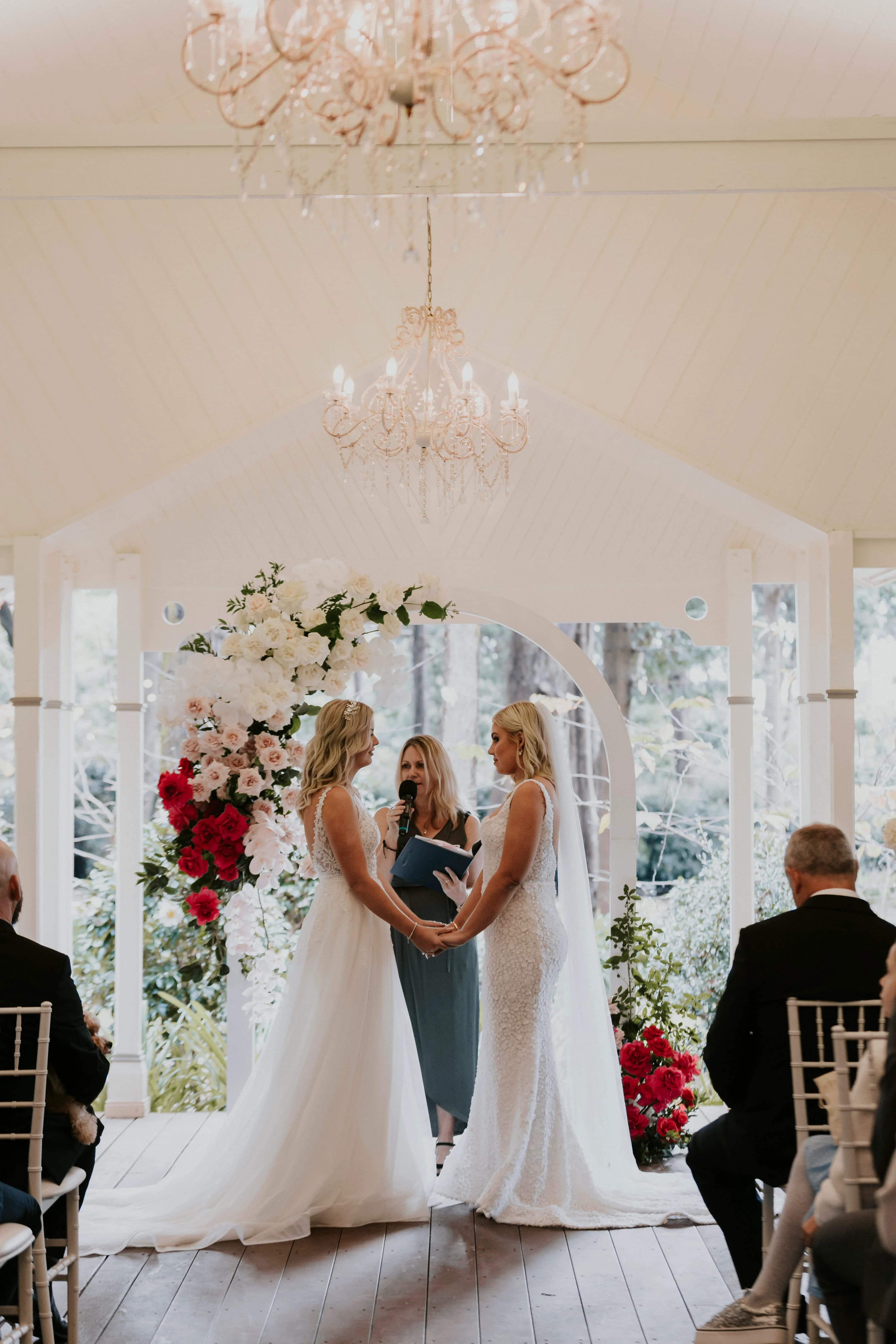 Two brides stand facing each other, holding hands under a floral arch adorned with white and red flowers. A celebrant in a blue dress stands between them, speaking into a microphone. Guests are seated on either side in a stylish, chandelier-lit venue.