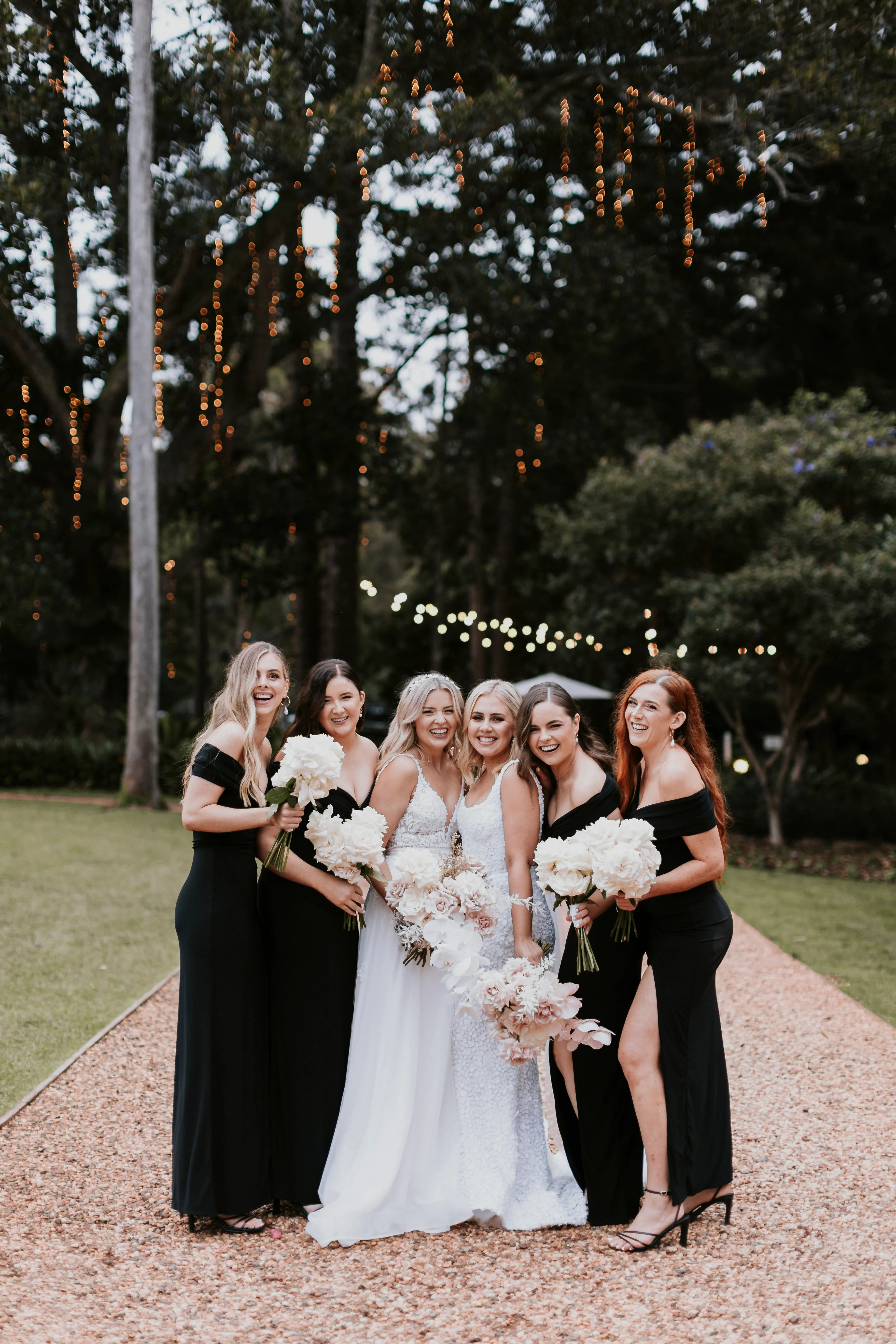 A bride in a white dress, holding white flowers, stands on a gravel path surrounded by five bridesmaids in black dresses, also holding white bouquets. They are outdoors with trees and string lights in the background, all smiling and posing for the photo.