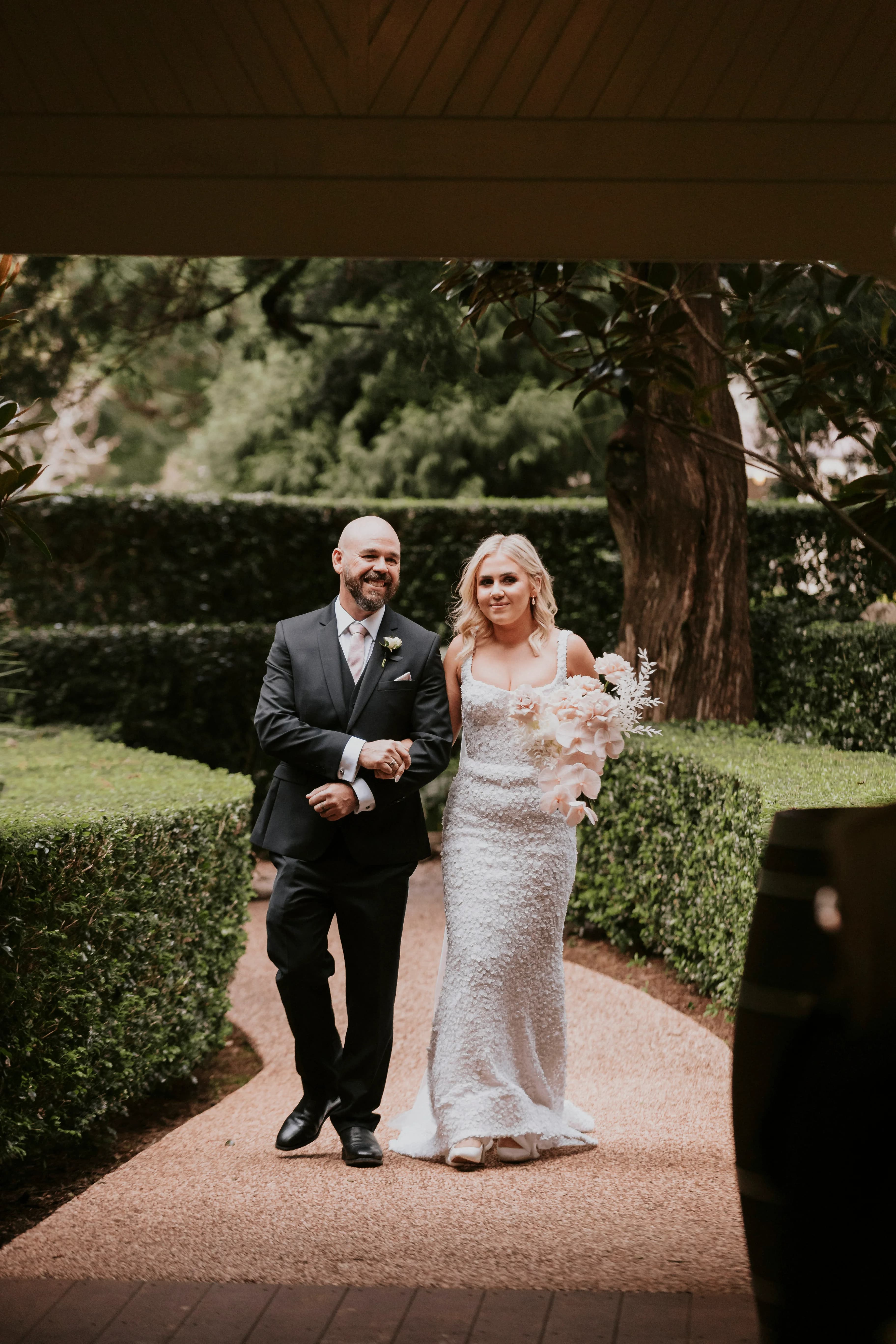 A bride, dressed in a white lace wedding gown and holding a bouquet of flowers, walks arm-in-arm with a man in a black suit and tie with a boutonniere. They are walking down a garden path with lush green hedges and trees in the background.