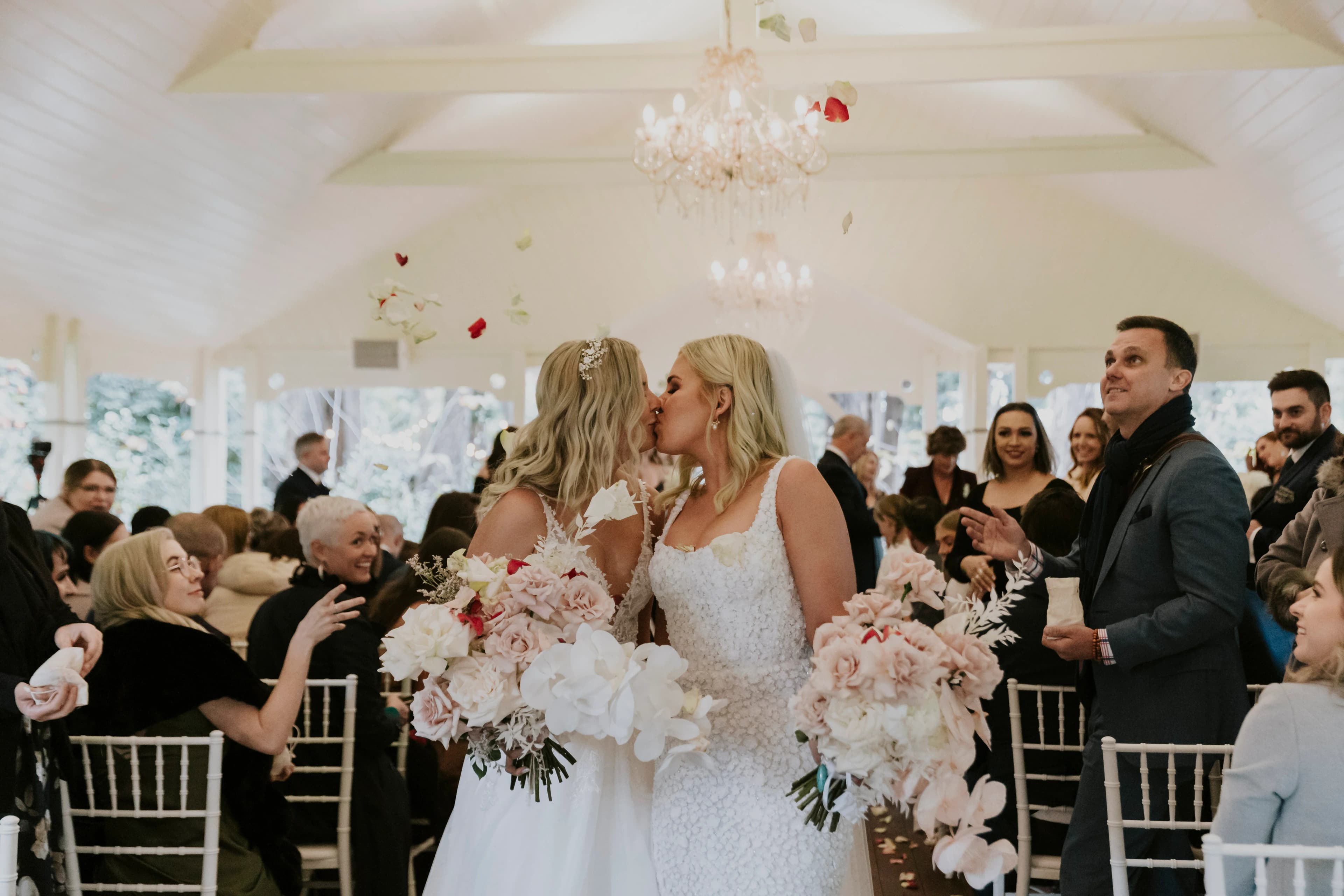 Two brides in white dresses kiss at the end of the aisle in a light-filled room as guests look on and smile. Both brides hold large bouquets of pink and white flowers. Flower petals float in the air above them. The room is decorated with chandeliers and white chairs.