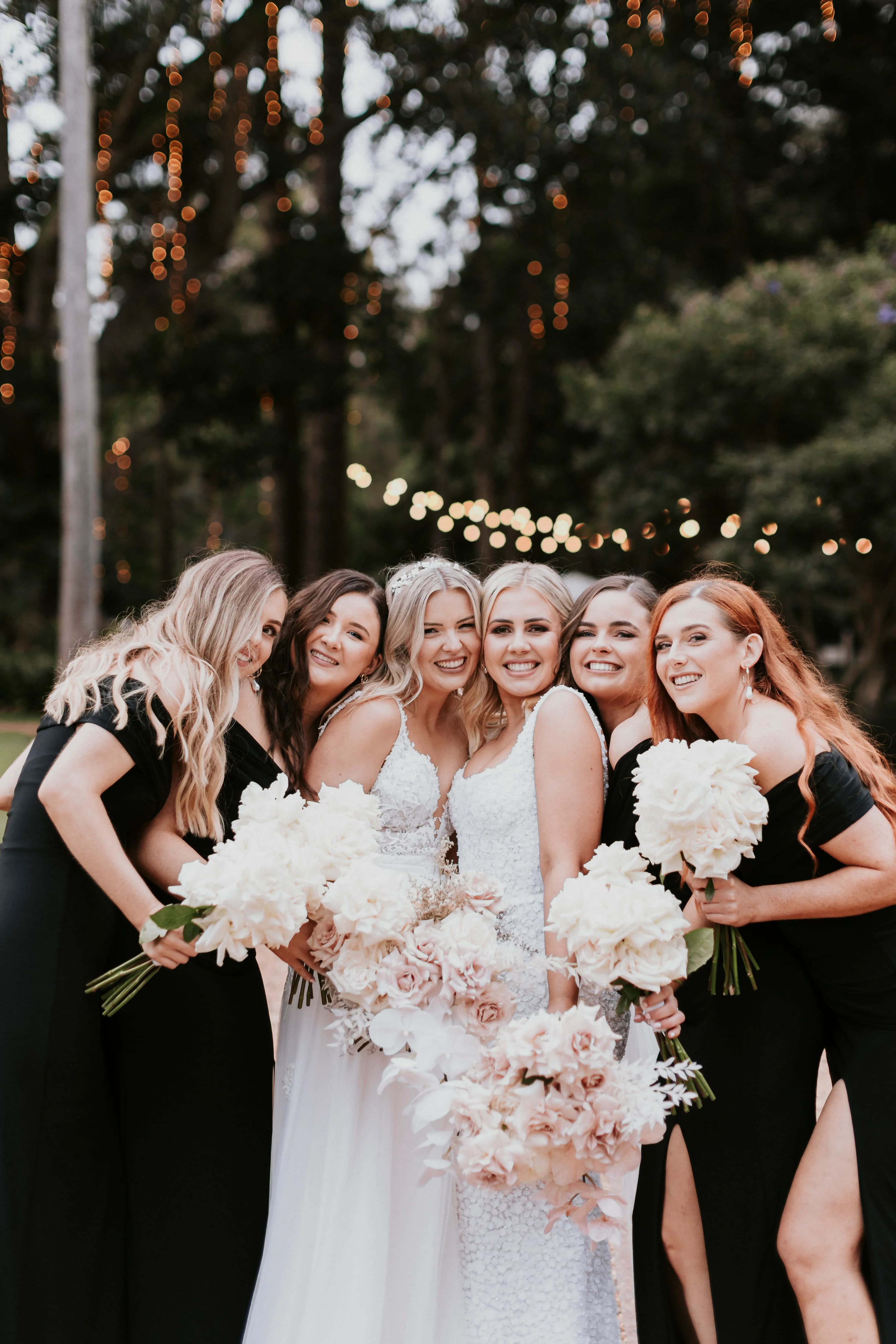 A group of six women wearing black dresses and holding white and pink bouquets smile and pose together in an outdoor setting. Two women in the middle wear white dresses, likely brides. String lights are visible in the background amidst trees, creating a festive atmosphere.