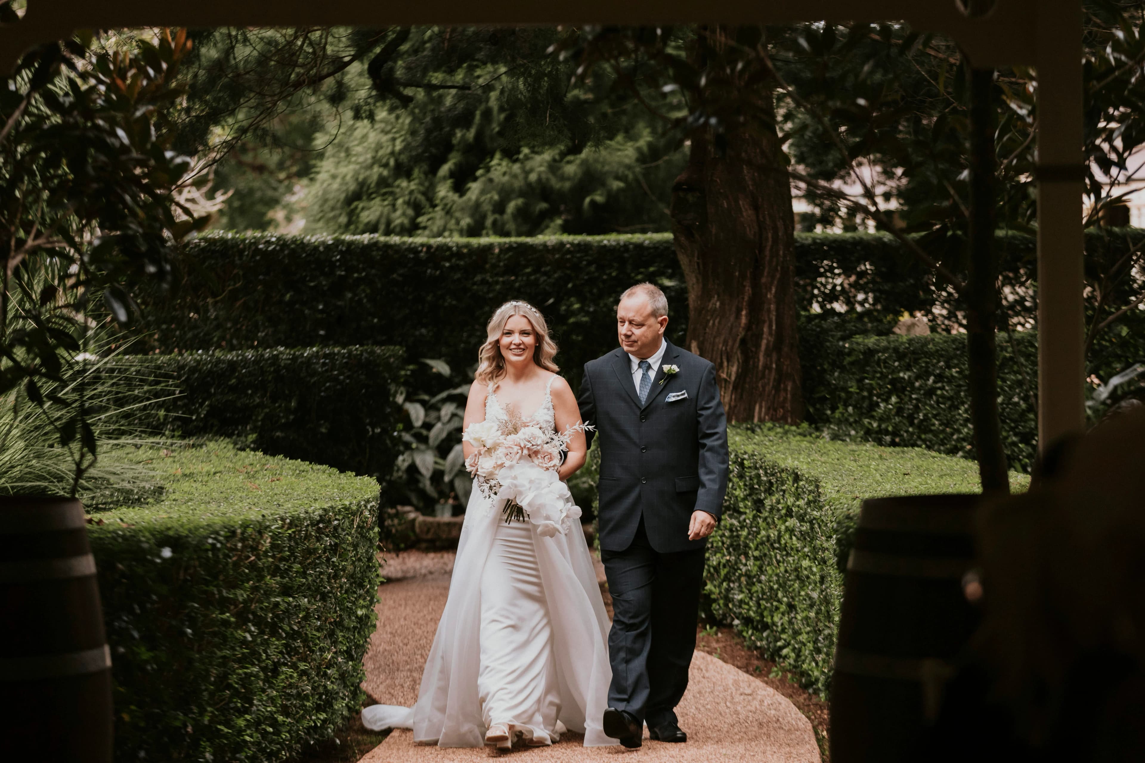 A bride in a white wedding dress holding a bouquet walks down a garden path with a man in a dark suit. They are surrounded by lush green hedges and trees, creating a serene and picturesque setting. Both are smiling and appear to be enjoying the moment.