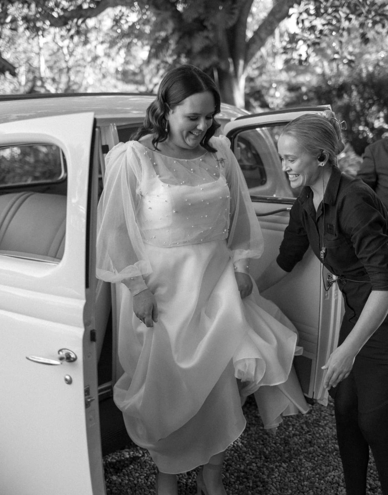 A black and white photo of a smiling bride stepping out of a vintage car, assisted by another woman. The bride wears a flowing, satin wedding gown with sheer sleeves and delicate beading, while the other woman, smiling too, wears a dark, formal outfit.