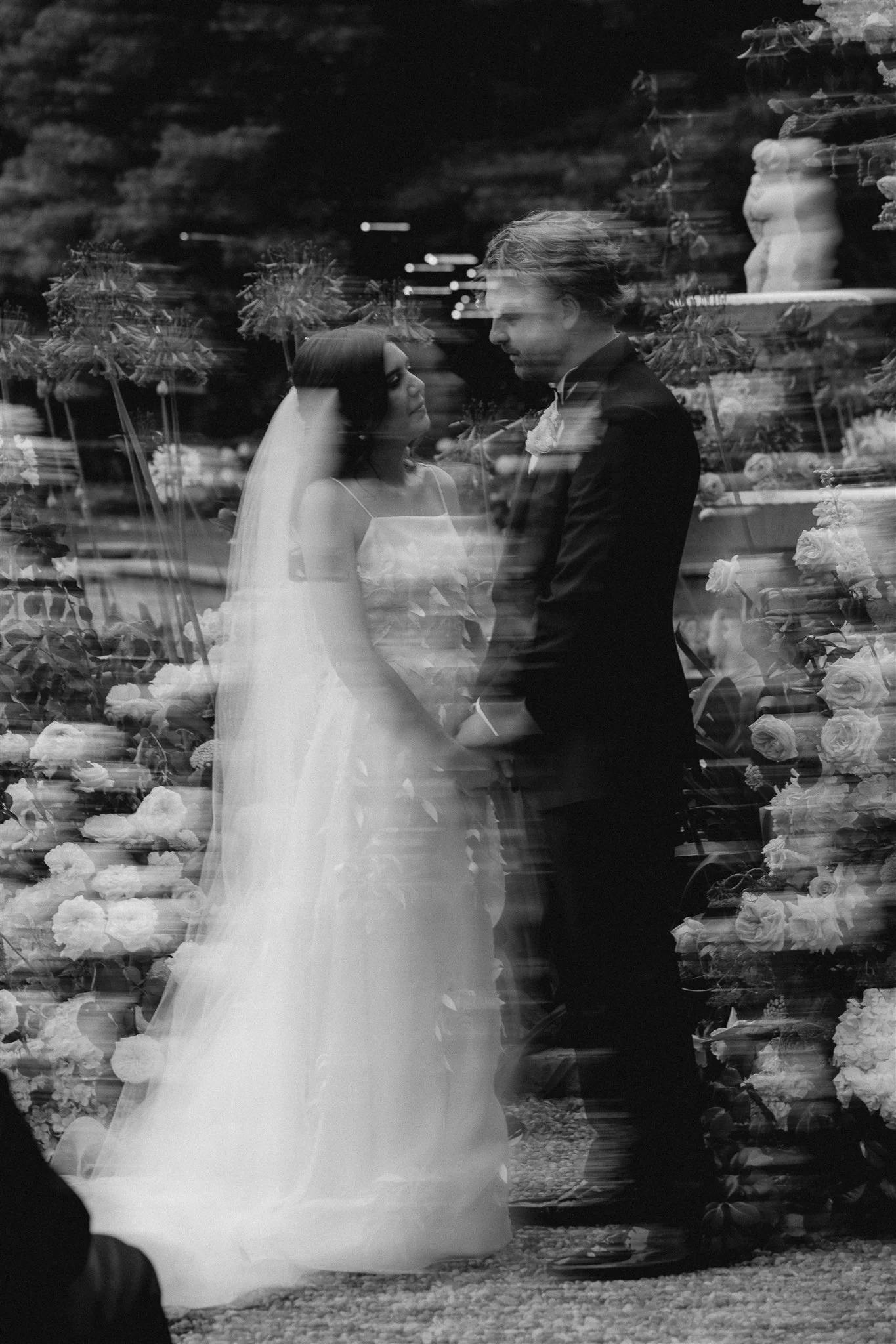 A black and white photo of a bride and groom standing close to each other, holding hands amidst a lush floral arrangement. The bride is wearing a flowing gown and a veil, while the groom is in a suit. The image has a dreamy, double exposure effect.