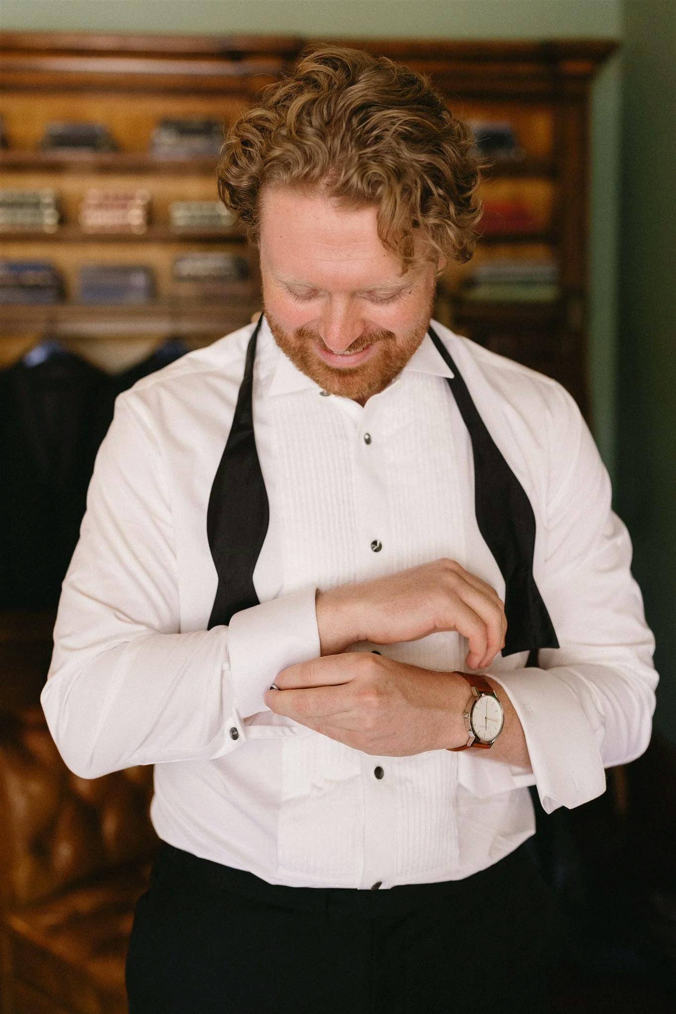 A man with curly hair and a beard adjusts the cuff of his white dress shirt. He is wearing a black bow tie that is untied around his neck. Behind him is a wooden shelf with neatly folded fabrics.