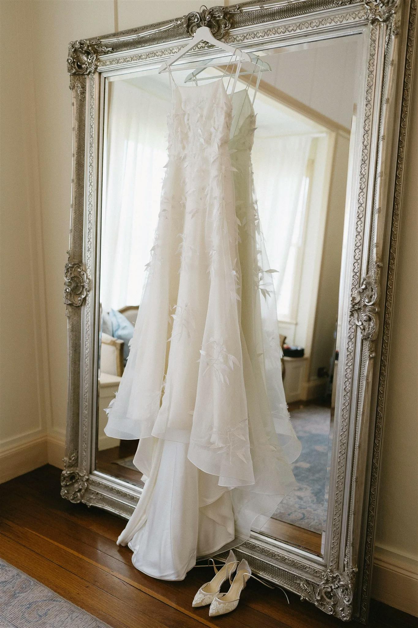 A white wedding dress with delicate floral appliqué details hangs on an ornate silver-framed mirror. Below the dress, on the wooden floor, a pair of white lace heels are placed neatly. The room features off-white walls and a glimpse of light curtains and furniture.