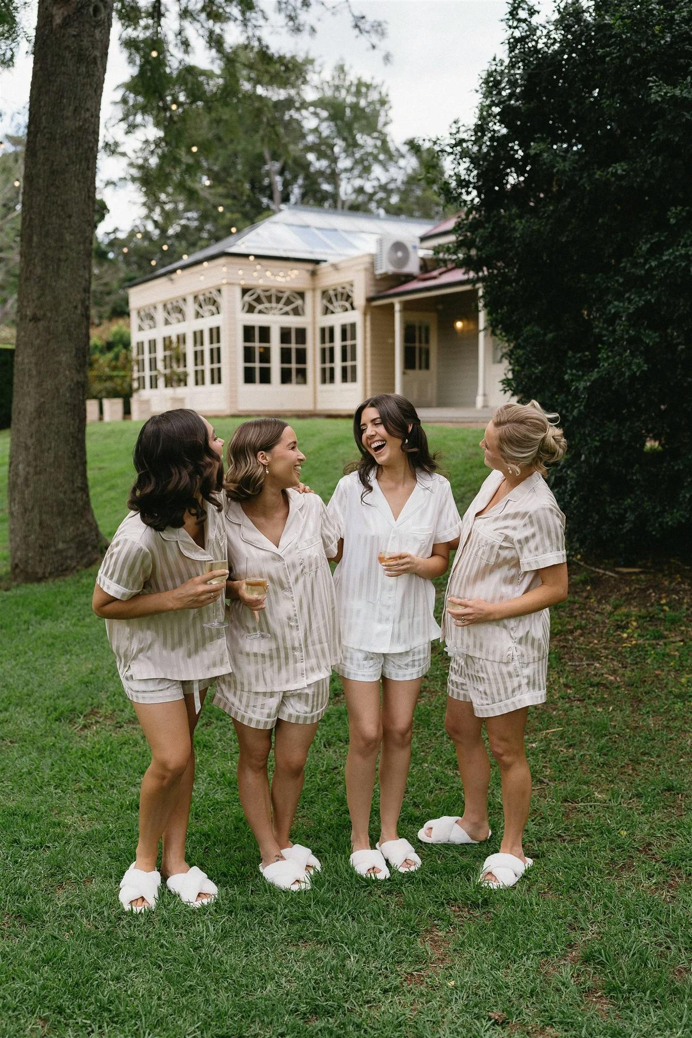 Four women wearing matching pajamas and slippers stand on a lawn, laughing and chatting. They are in front of a large building with a glass-paned facade and surrounded by trees and greenery.