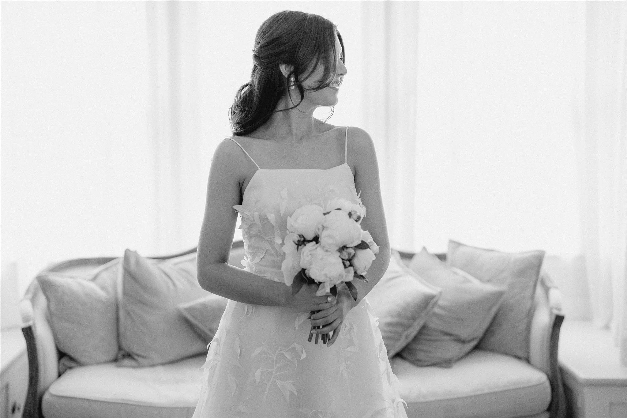 A bride in a white dress stands with a bouquet of flowers. The photo is in black and white, with soft curtains in the background and a couch with cushions behind her. She is looking to the side, giving the image a classic and elegant feel.