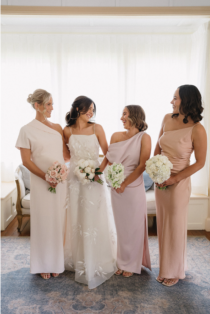 A bride in a white floral gown stands with three bridesmaids in pastel dresses. Each holds a bouquet and smiles while looking at one another. They are in a well-lit room with large windows and a patterned rug.