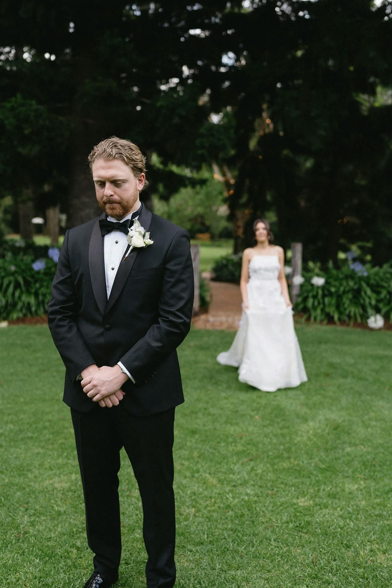 A bride in a white wedding dress stands a few feet behind a groom in a black tuxedo, who has his eyes closed and hands clasped in front of him. They are outdoors on a lawn with greenery and flowers in the background.