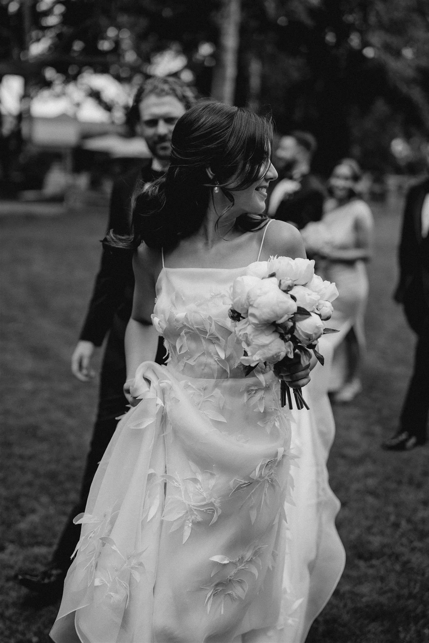 A bride in a white floral-patterned wedding gown holds a bouquet of flowers and smiles while looking to her side. She is surrounded by people, including a man in a suit walking beside her. The background shows a grassy area with trees. The image is in black and white.