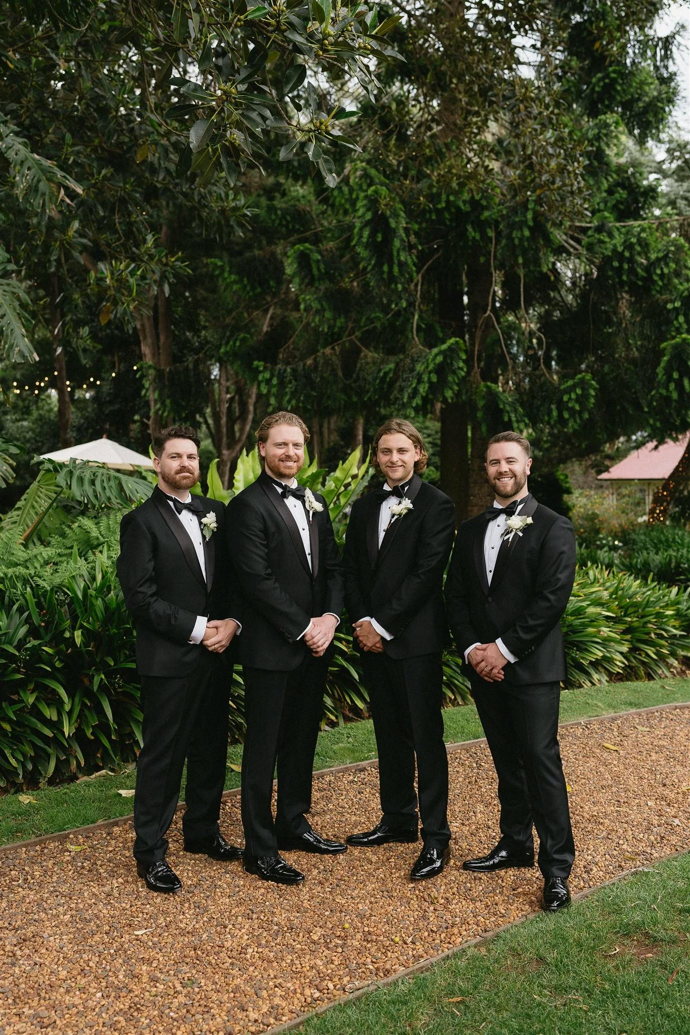 Four men in black tuxedos stand on a gravel path in an outdoor garden. Trees and lush greenery surround them, and they each have a boutonniere pinned to their lapels. They are smiling and posing for the photo.