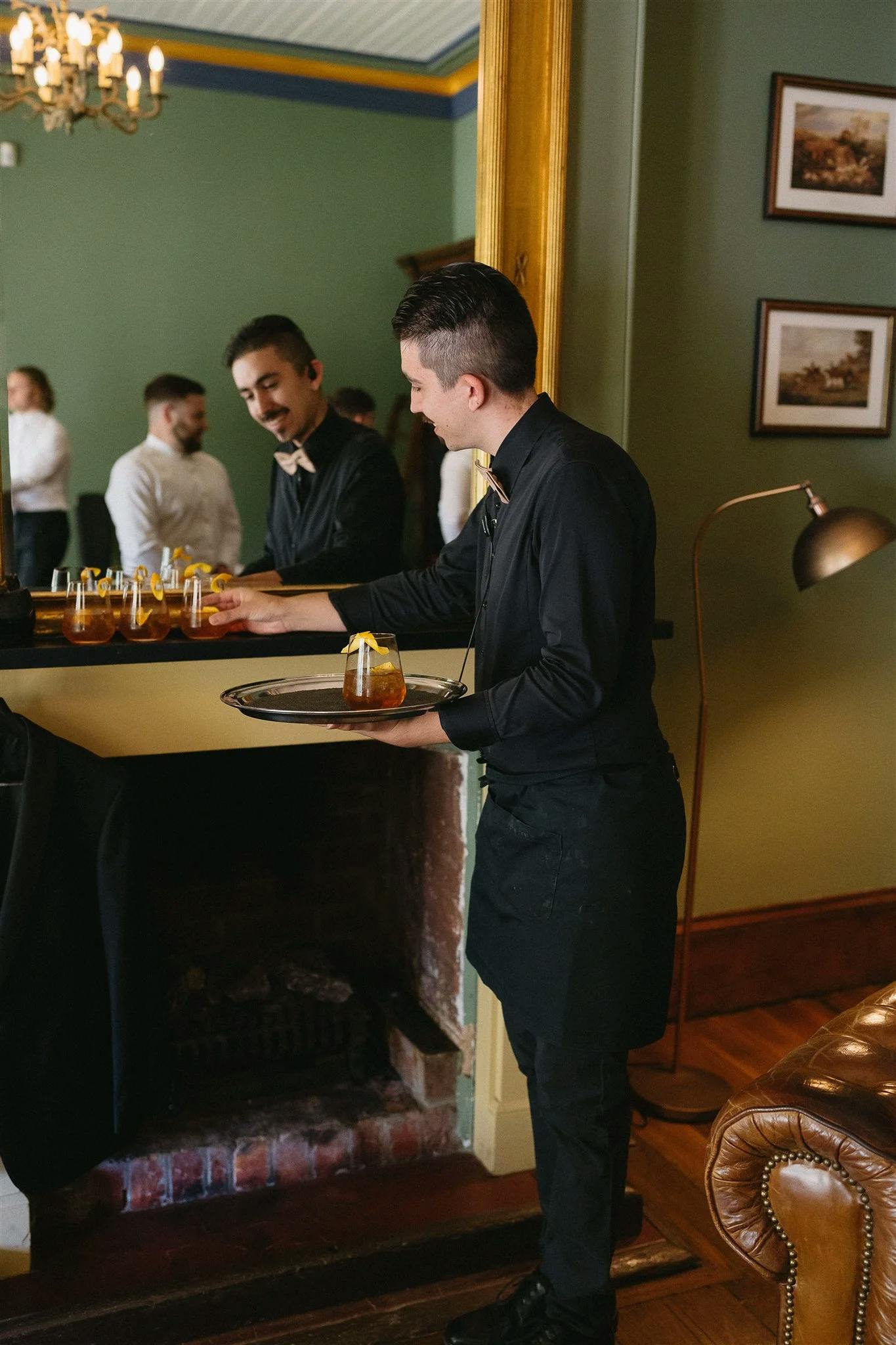 A waiter in a black uniform and apron serves a cocktail from a tray in a dimly lit, elegant room with green walls. A man in a white shirt and tie stands near a polished black counter holding drinks. Pictures and a lamp adorn the room, and another person is visible in the background.